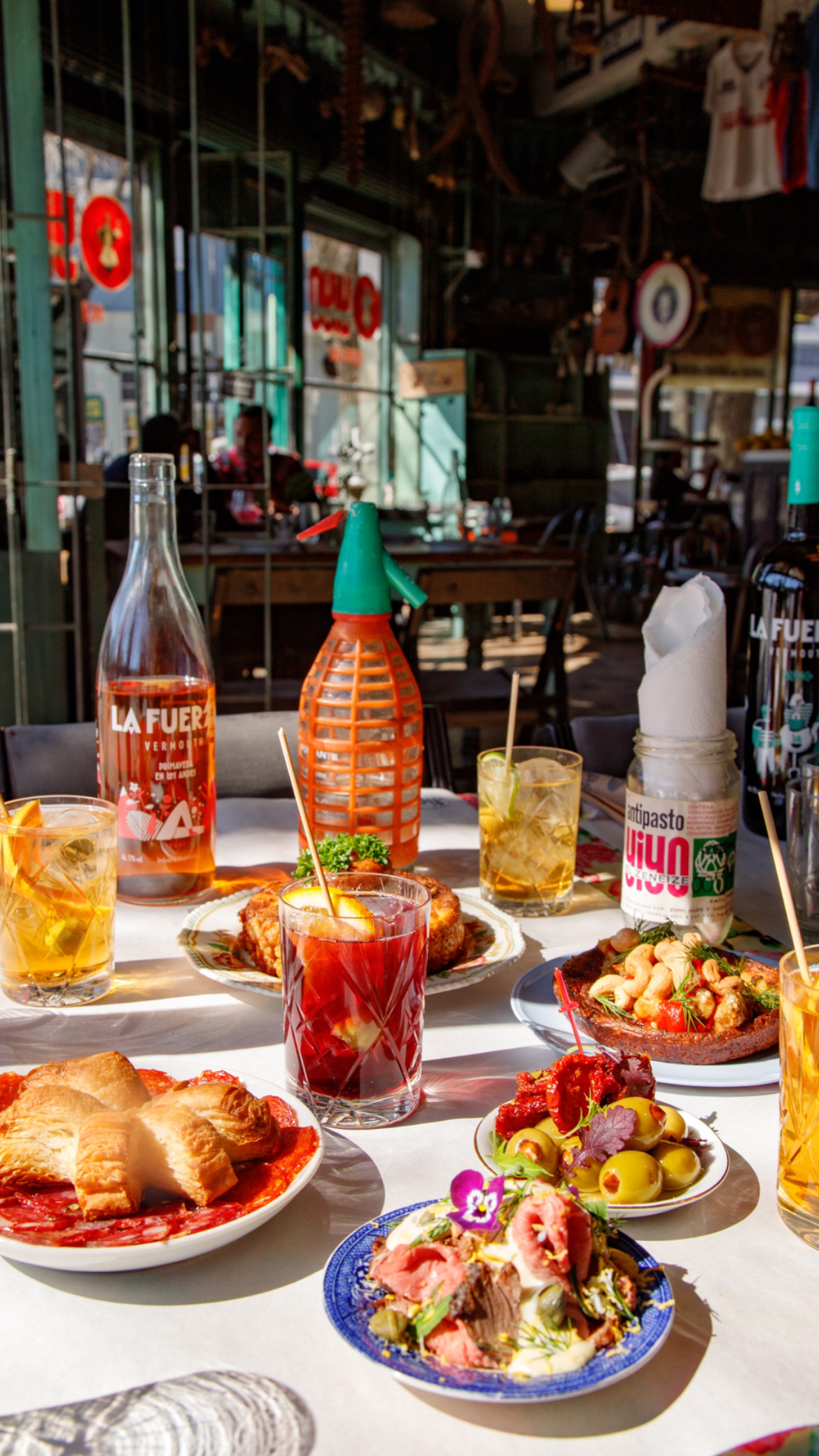 A table filled with bright colorful dishes, a red cocktail, bottles of aged amaro and seltzer