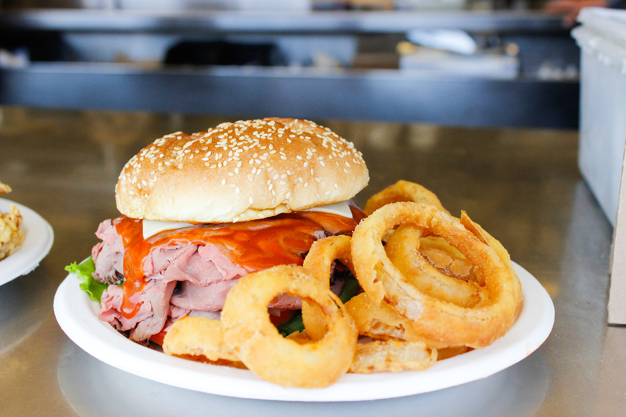 A roast beef sandwich with cheese, mayo, and barbecue sauce sits on a paper plate, accompanied by thick onion rings.