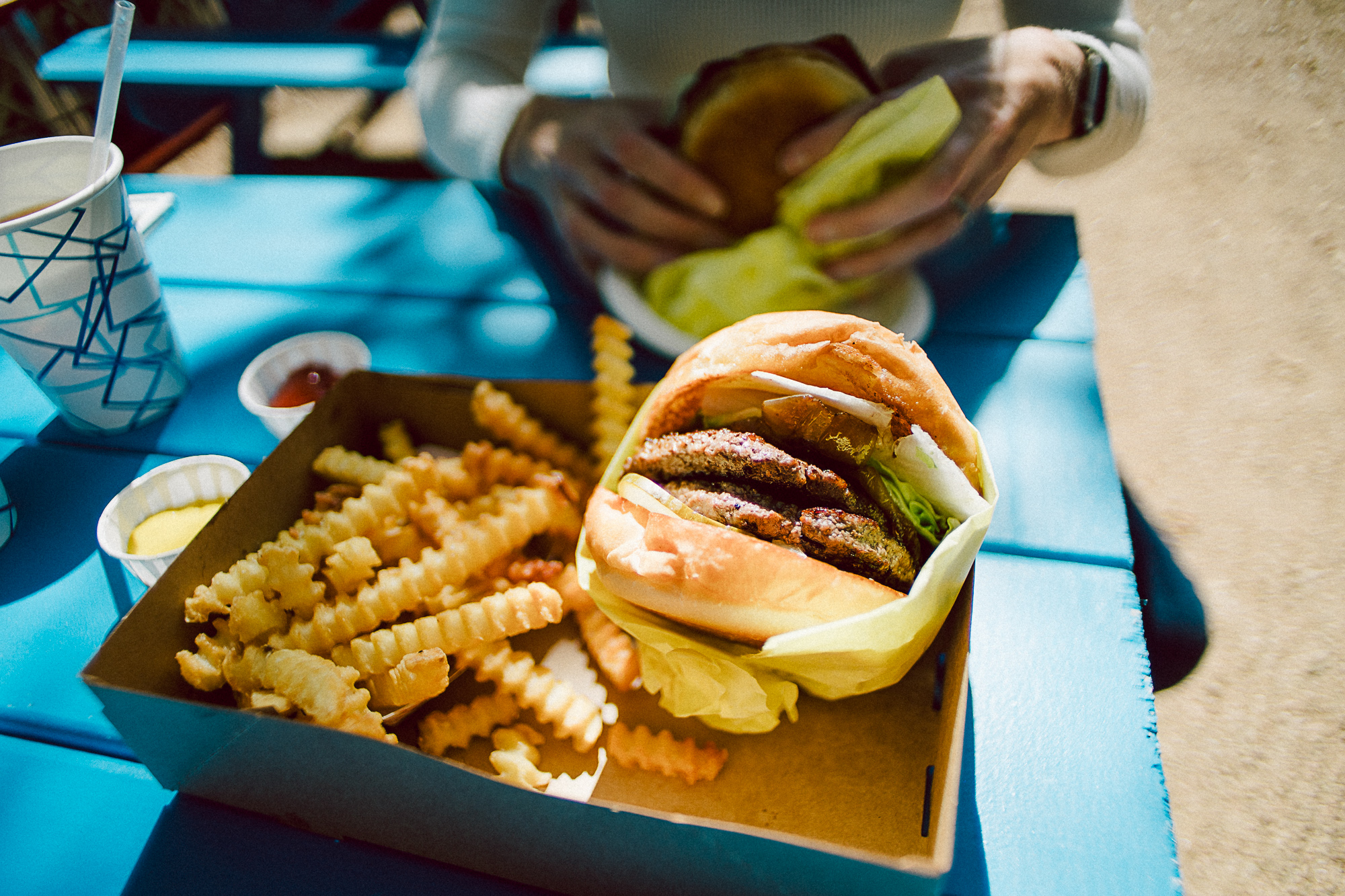 A burger in paper wrap and fries in a tray on a blue picnic table.