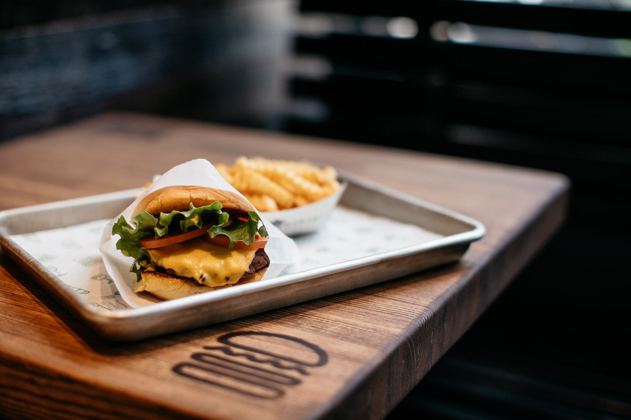 A shack burger on a metal tray on a wood table next to a basket of crinkle cut fries.