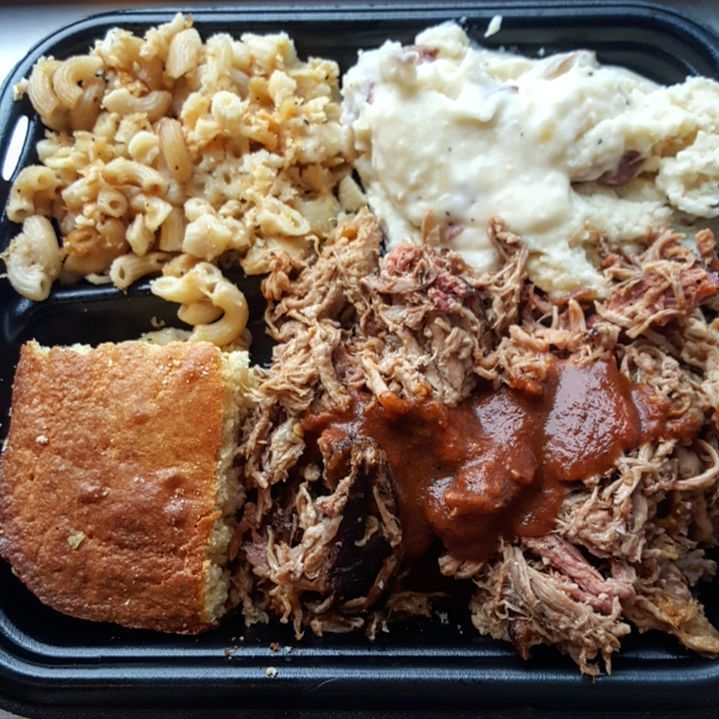 Overhead closeup view of a black plastic takeout container stuffed with mac and cheese, mashed potatoes, pulled pork with barbecue sauce, and cornbread