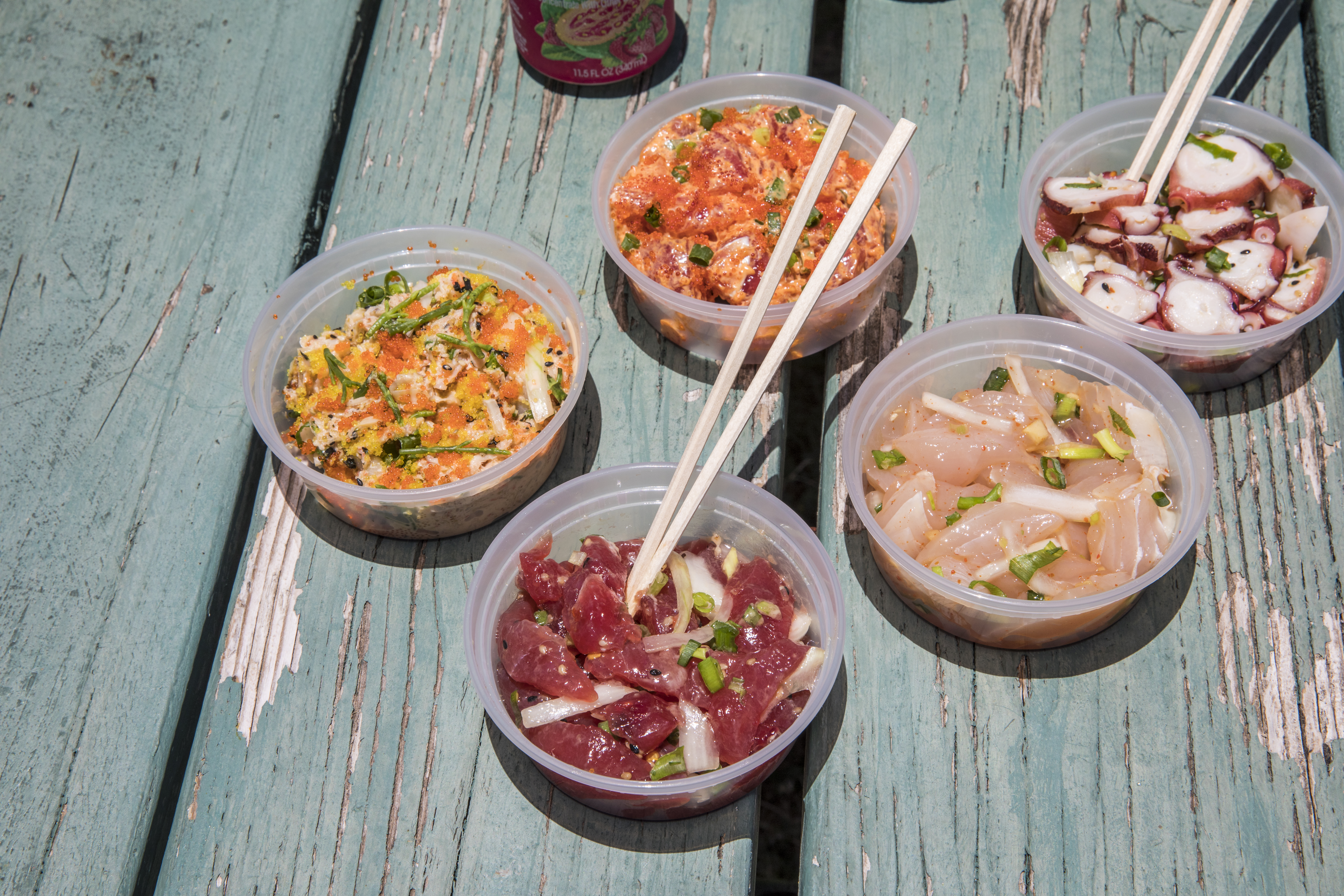 Various kinds of poke in plastic containers with drinks and chopsticks on a light blue picnic table.
