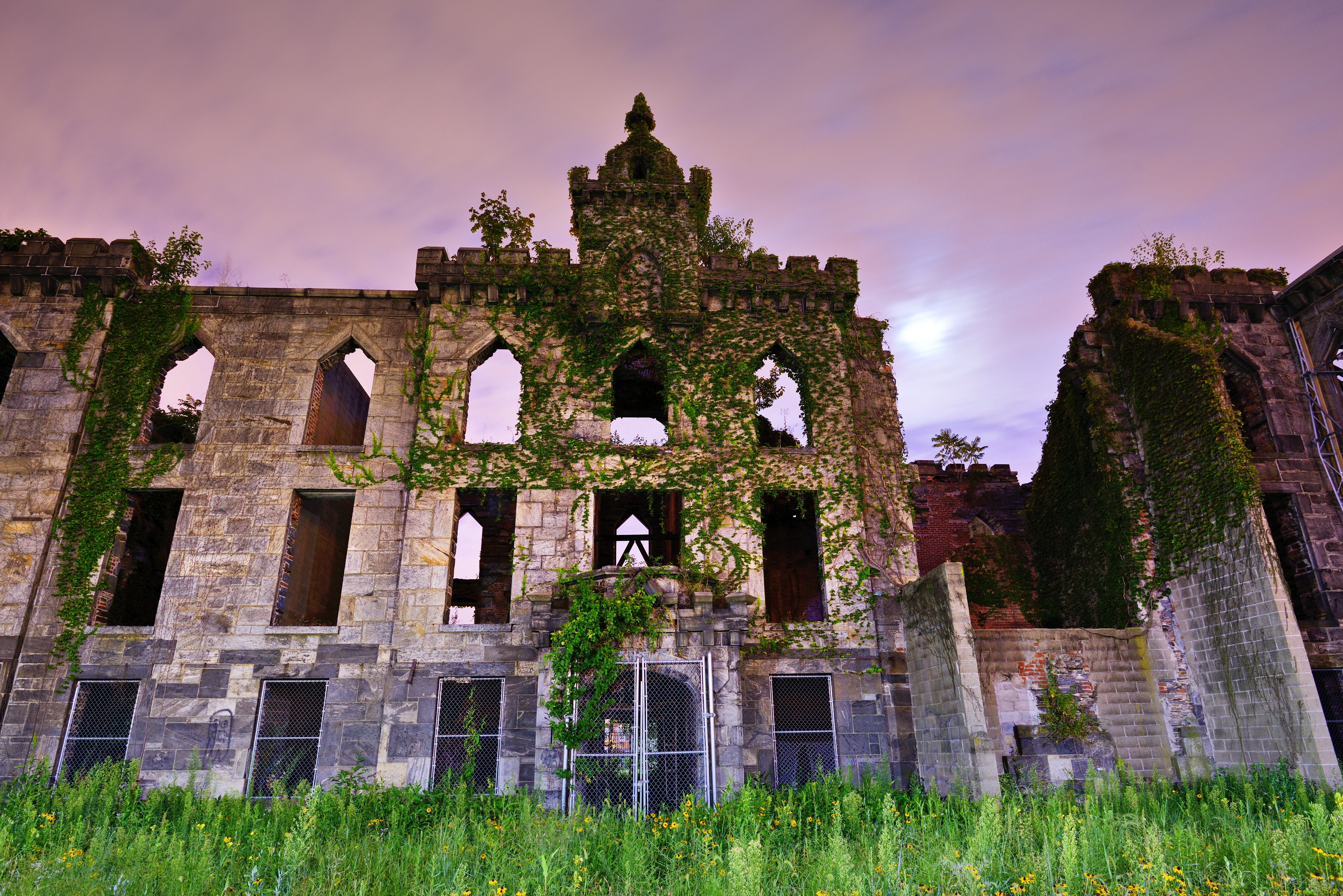 The exterior of the Renwick Smallpox Hospital. The building is abandoned. There are multiple windows and the facade is stone. There is moss growing on some parts of the facade. It is evening and the sky is purple.