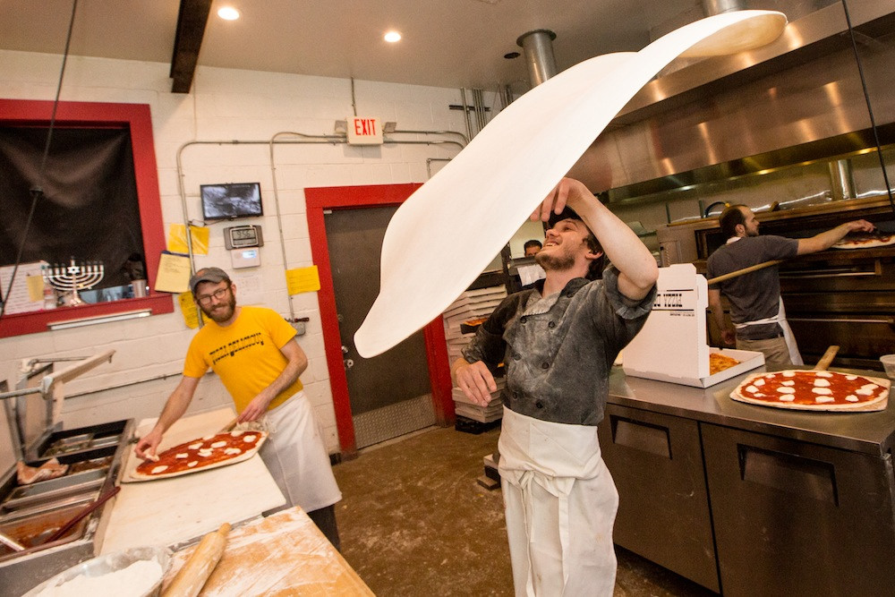 A person wearing a grey shirt and a white apron throwing a large round of pizza dough in a kitchen, with another person smiling in the background.