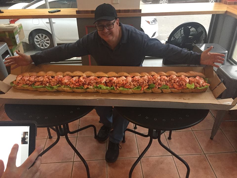 A man stands behind a giant five-foot-long lobster roll, arms outstretched to show off the length of the sandwich. It is sitting on a giant piece of cardboard spread across two small round tables.