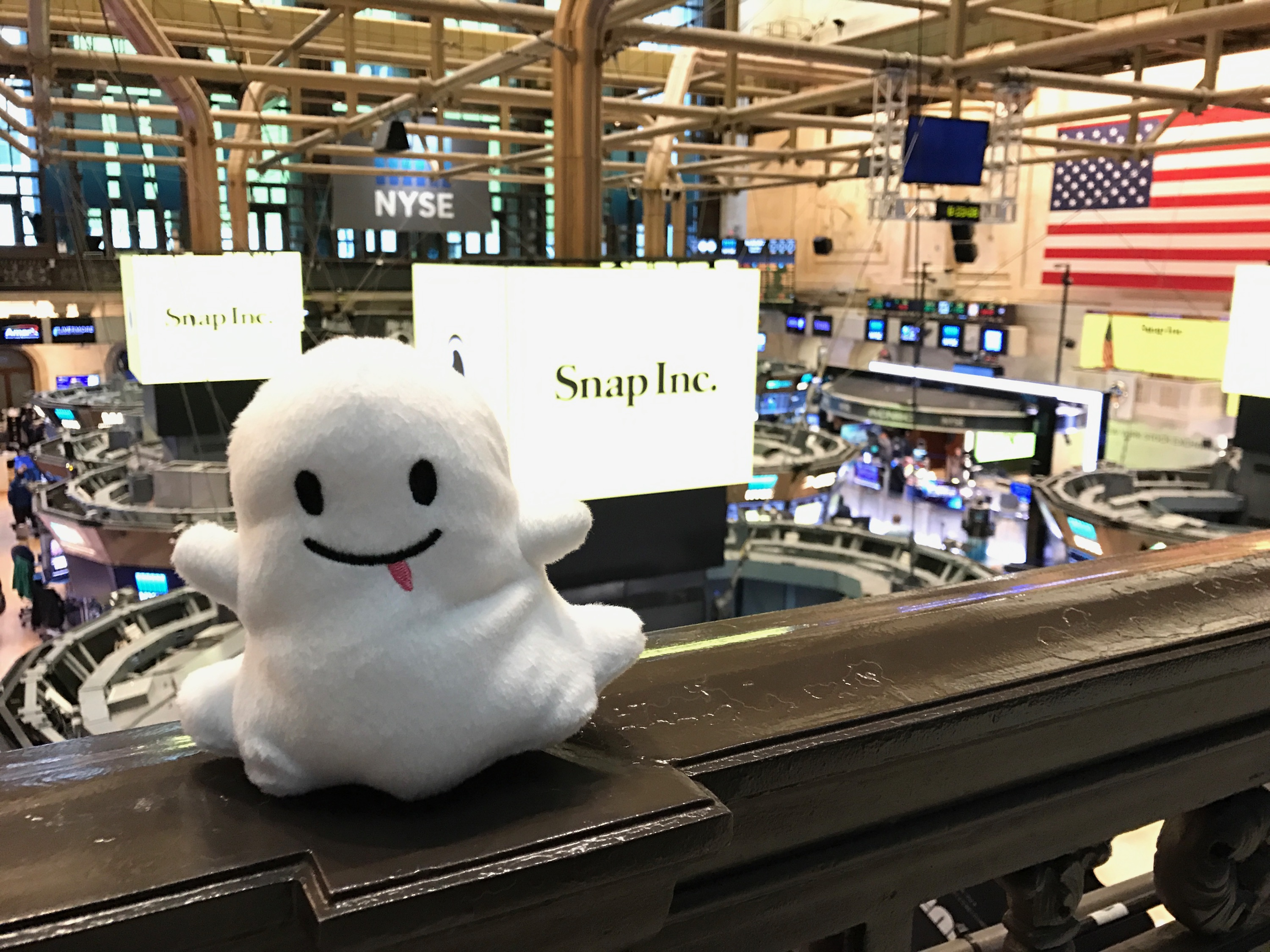 A stuffed and smiling ghost — the symbol for Snapchat — sits on a railing with the New York Stock Exchange in the background.
