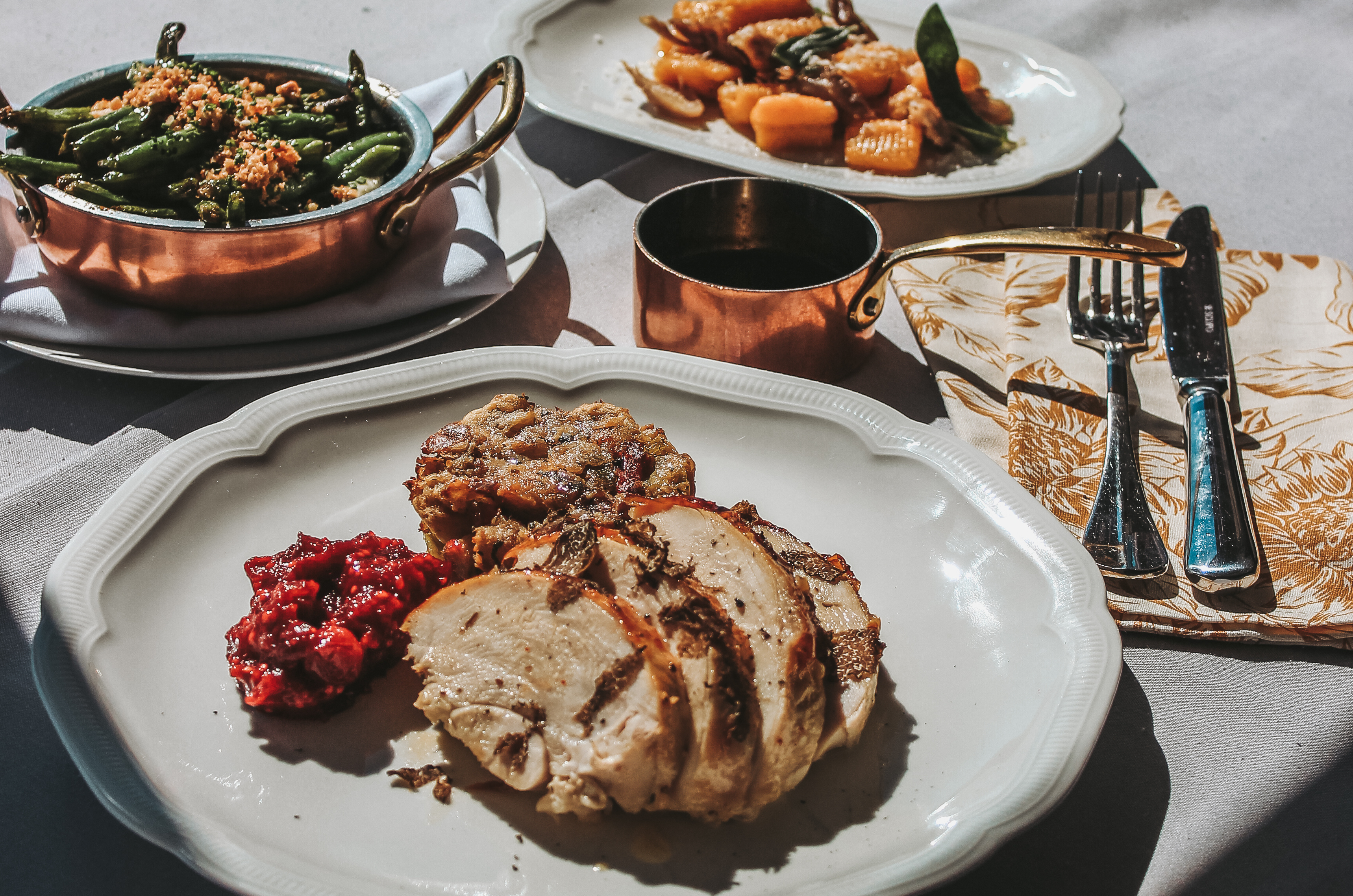 A table is covered with a white tablecloth. It holds plates of turkey with cranberry sauce, another with sweet potatoes and sage, and a third with a copper pan of green beans.
