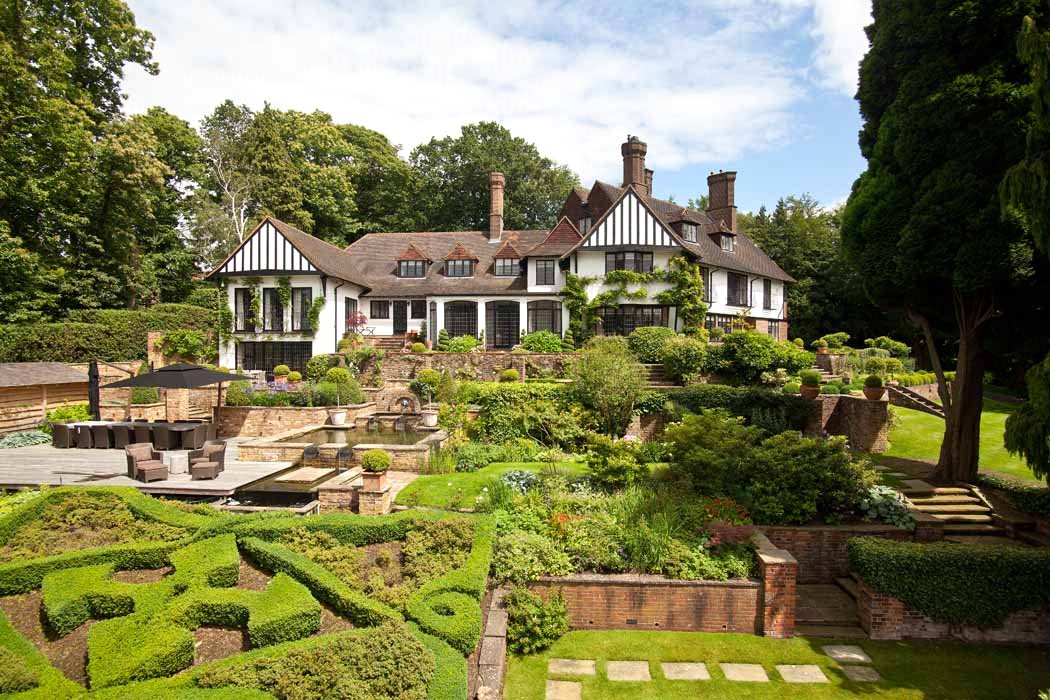 Exterior shot of Tudor-style house with multiple dormer windows and landscaped garden with fountain and hedges.