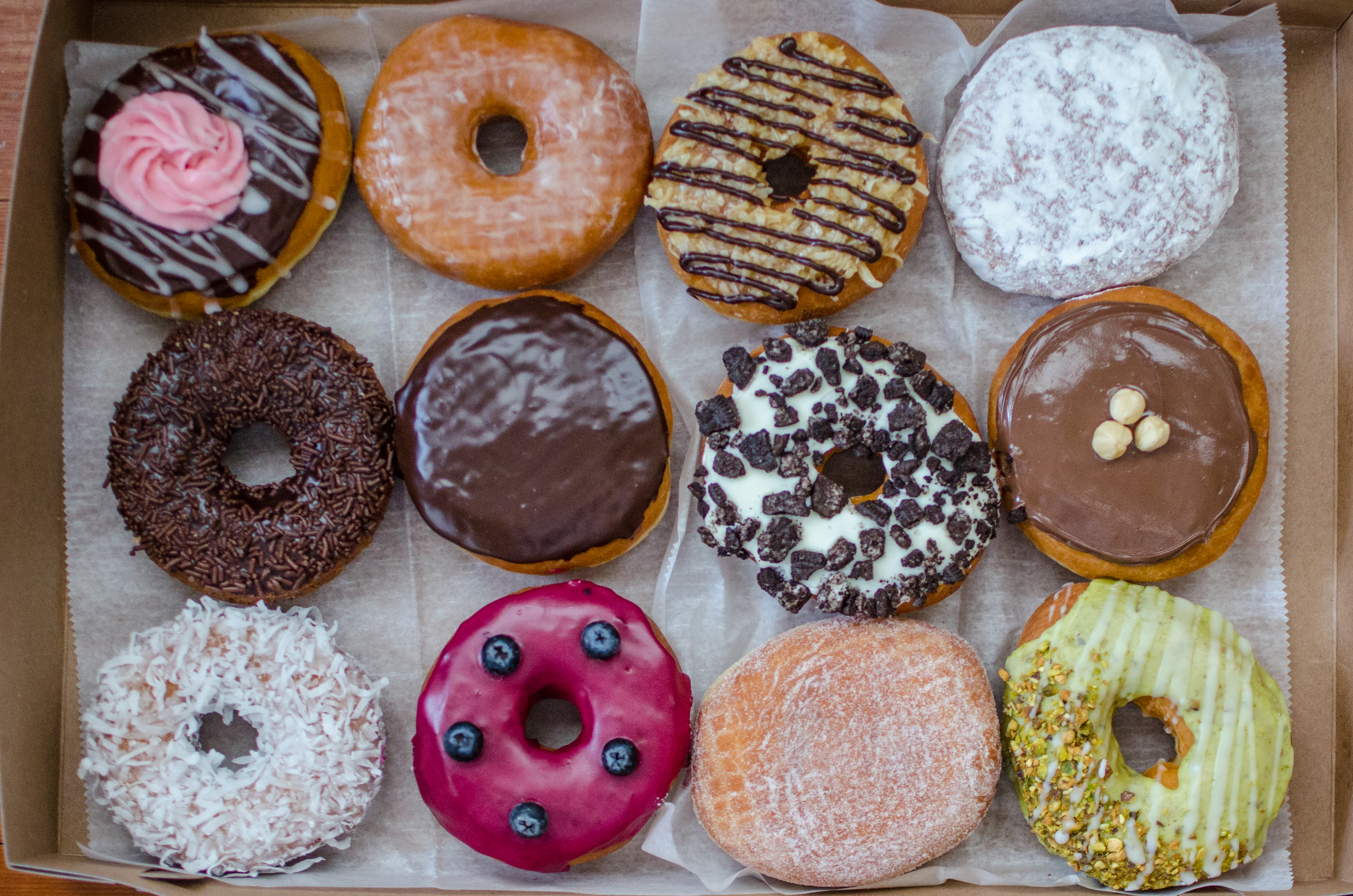 Overhead view of a dozen doughnuts, each a different flavor, lined up in three rows of four on wax paper in a cardboard box.