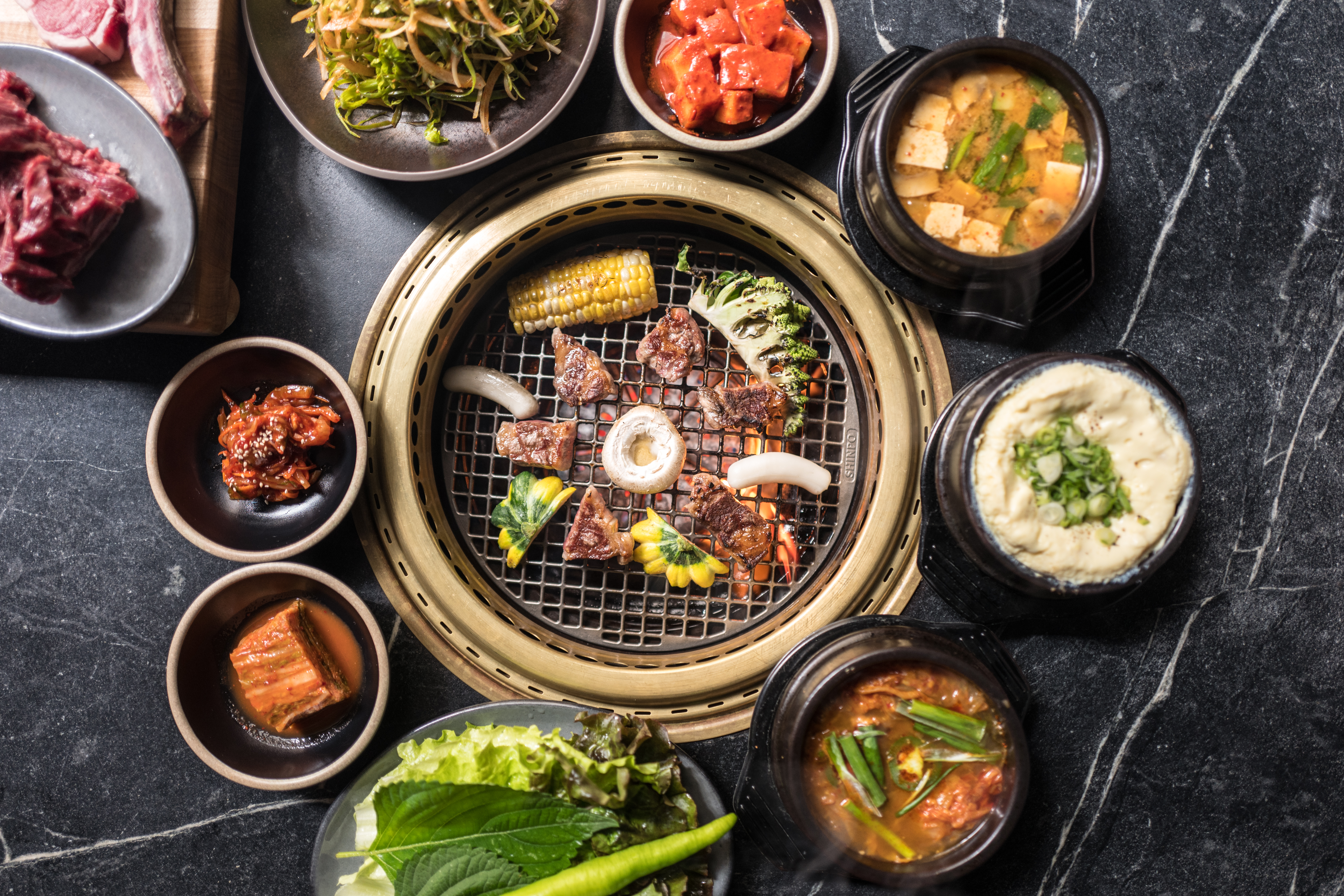 A circular beef-filled tabletop grill sits at the center; around that gold-rimmed grill are small banchan, including kimchi and egg omelet
