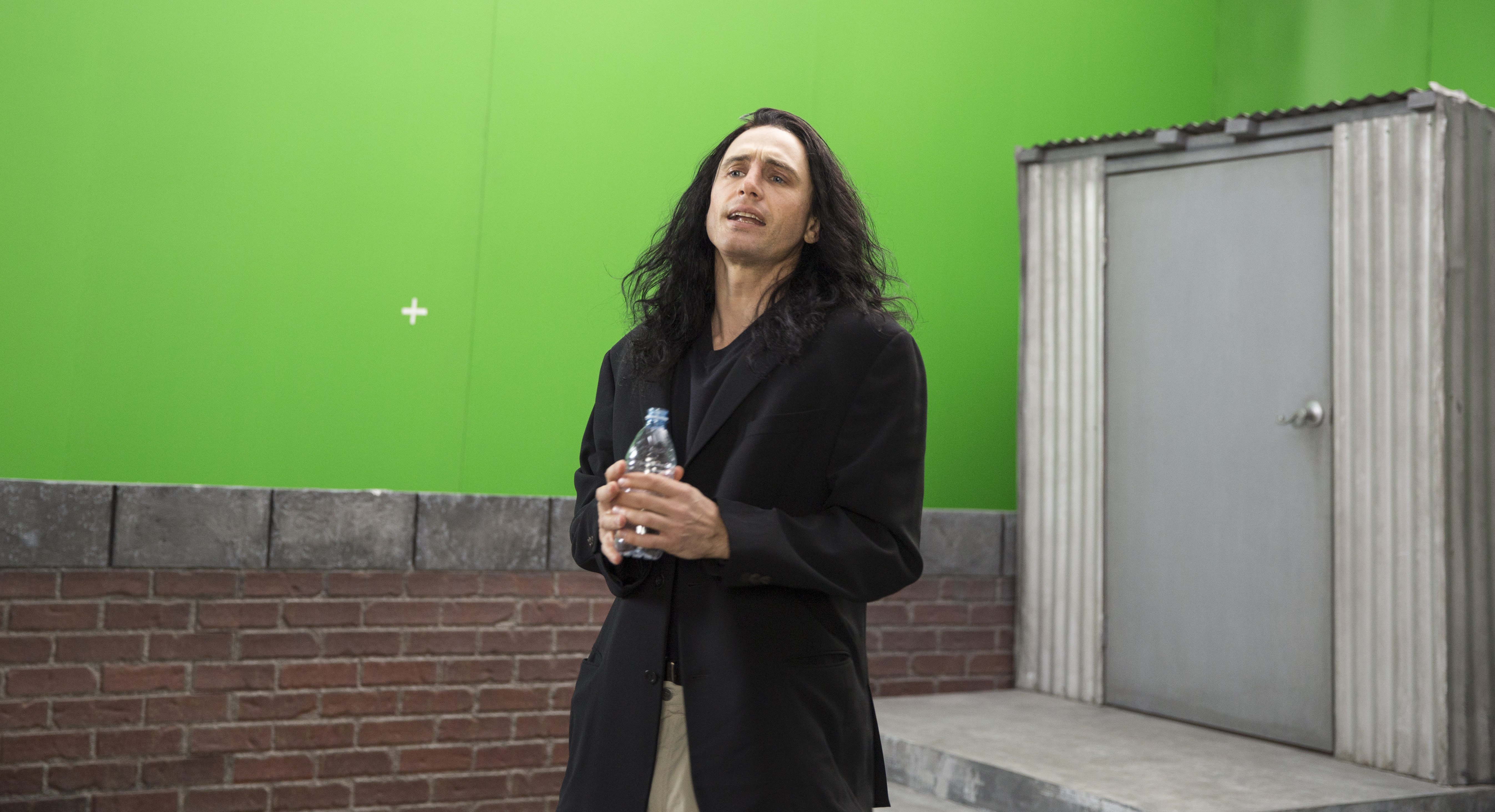 a man in a too big black jacket with long black hair in front of a green screen on a fake roof