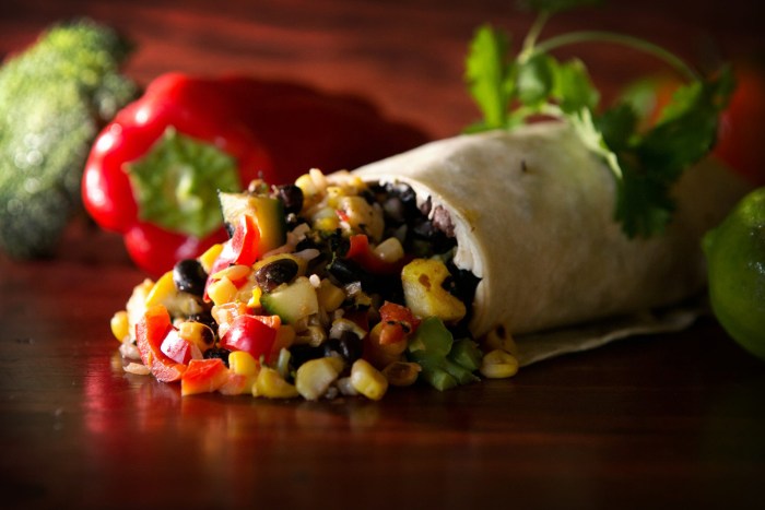 Corn, black beans, and red pepper pieces spill out of burrito on a dark wood surface. A red pepper and cilantro sit in the background.