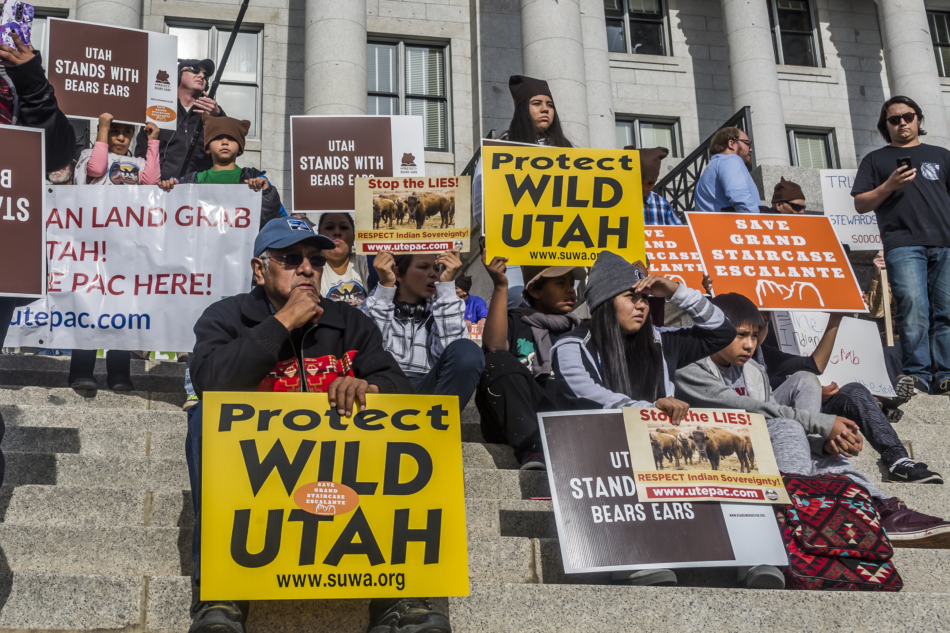 People sit on the capital building’s stone steps holding signs saying “Protect Wild Utah” and “Utah Stands With Bears Ears.”