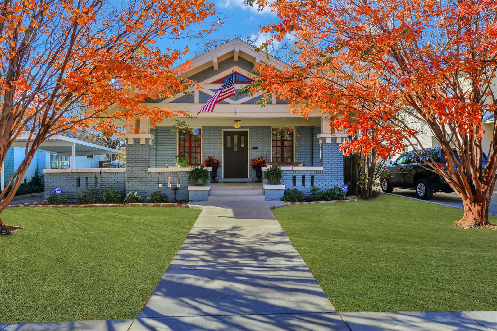 Exterior shot of blue-brick Craftstman style home with large front porch and a path leading to the house from the yard. Red-leaved trees frame the view. 