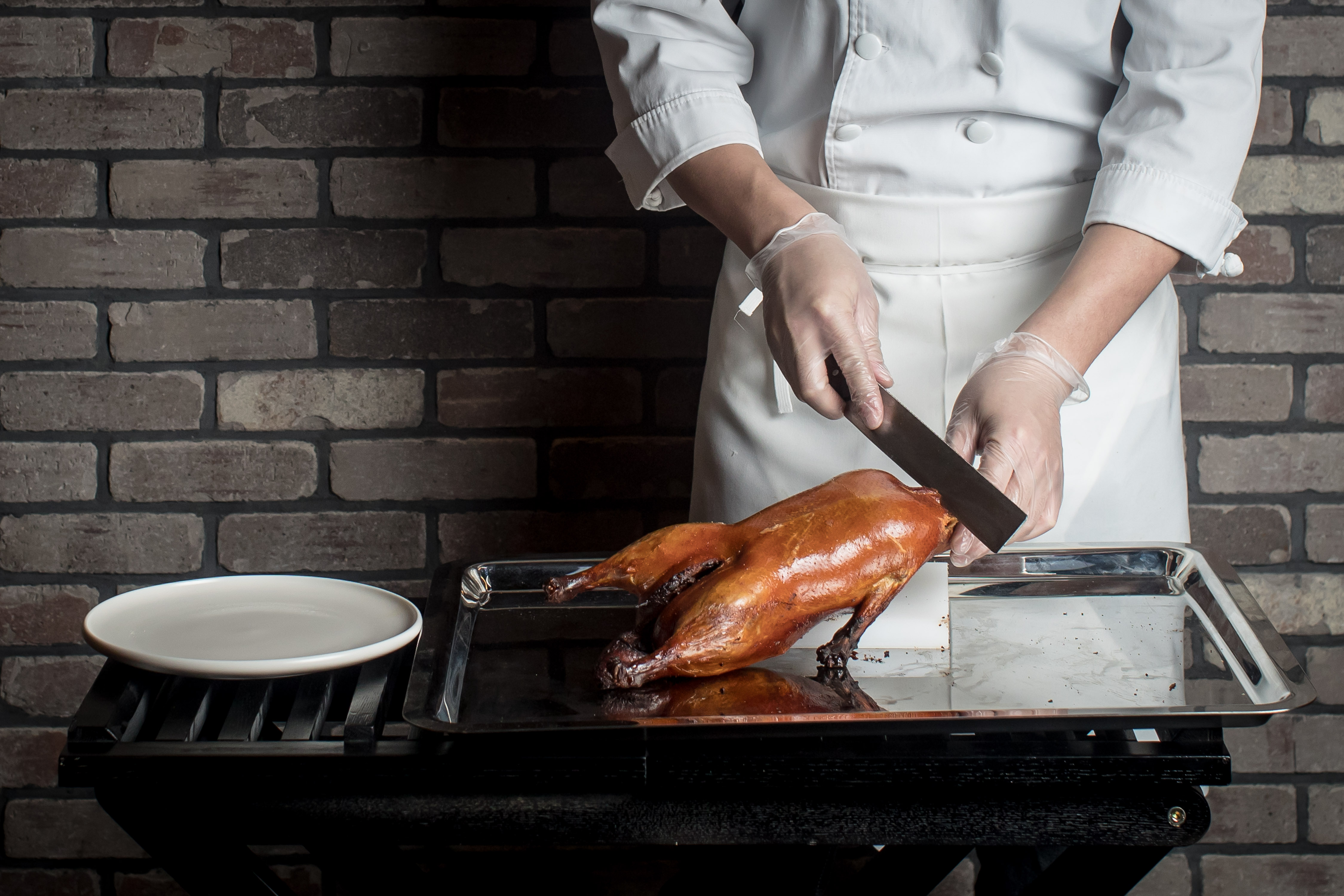 A chef carving a roasted duck with a flat-shaped knife. The duck is placed on a silver tray, and a white plate sits next to it.