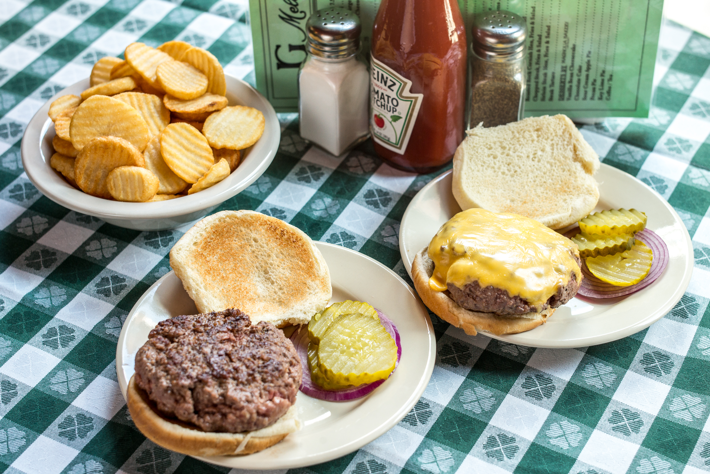 A burger and a cheeseburger at J.G. Melon sit on a green checkered tablecloth, while a plate of fries sits nearby.