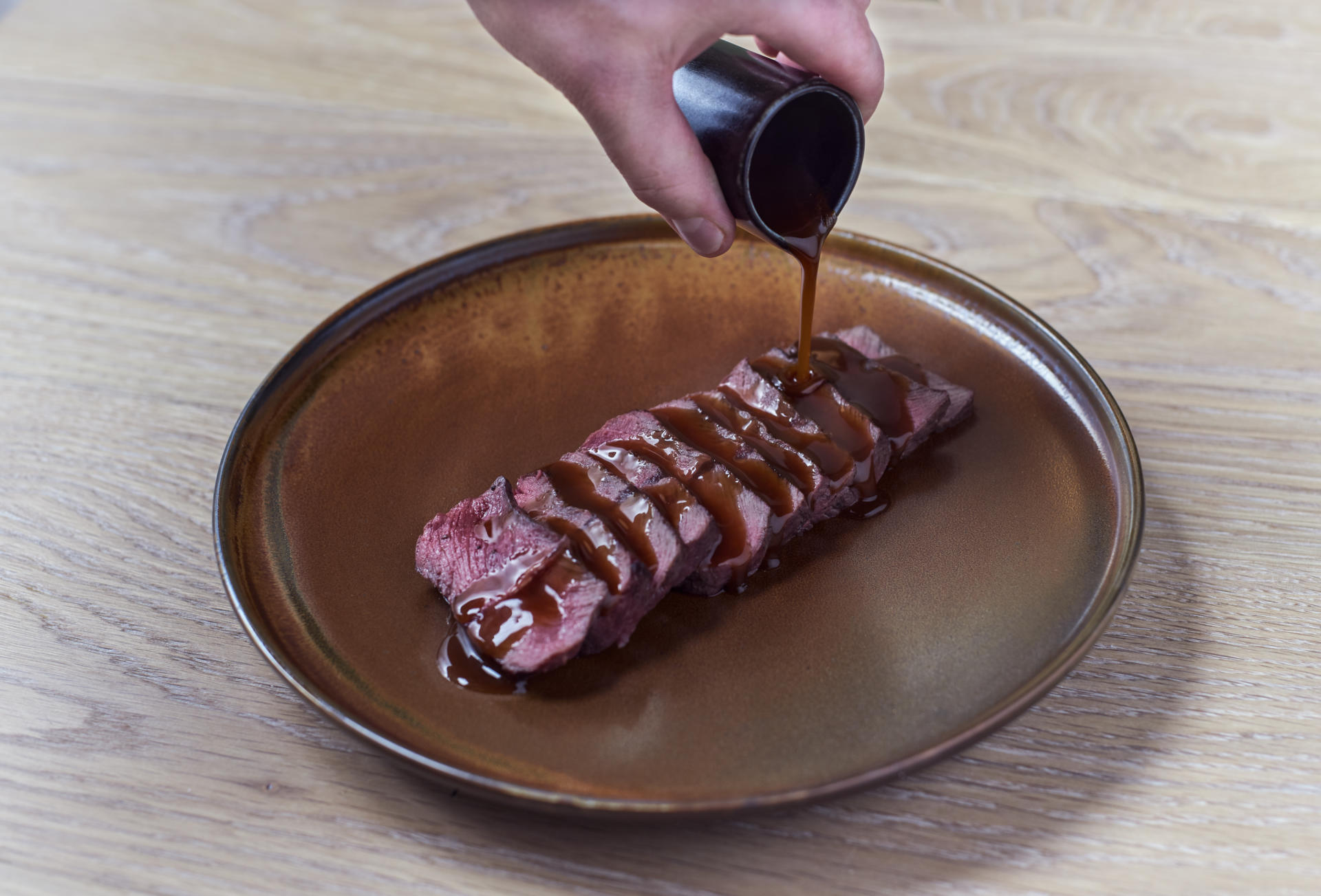 The hand of a server pours steak sauce over slices of beef arrayed on a decorative plate on a neutral wood background