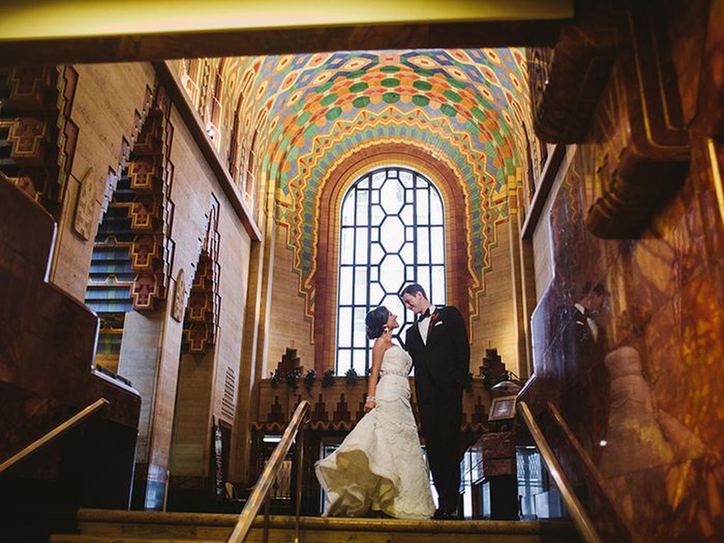 A woman and man embrace on the stairs in the Guardian Building in Detroit. The woman is wearing a wedding dress and the man is wearing a tuxedo. The ceiling is arched and decorated with an inlaid design.