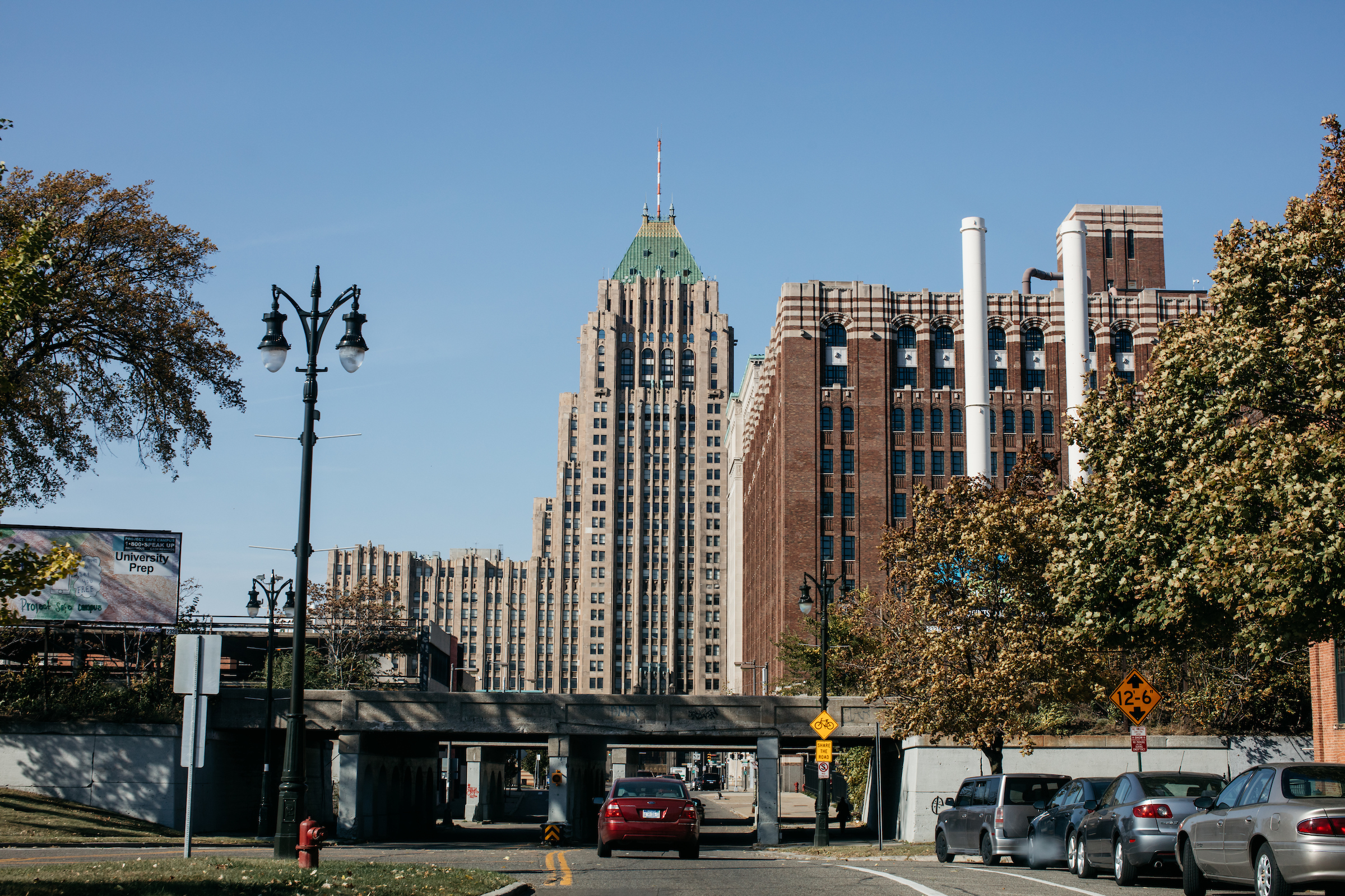 A road leads through an underpass in the foreground. In the background, there’s a tall brick building with white brick stripes and an even taller stone building with two towers. 