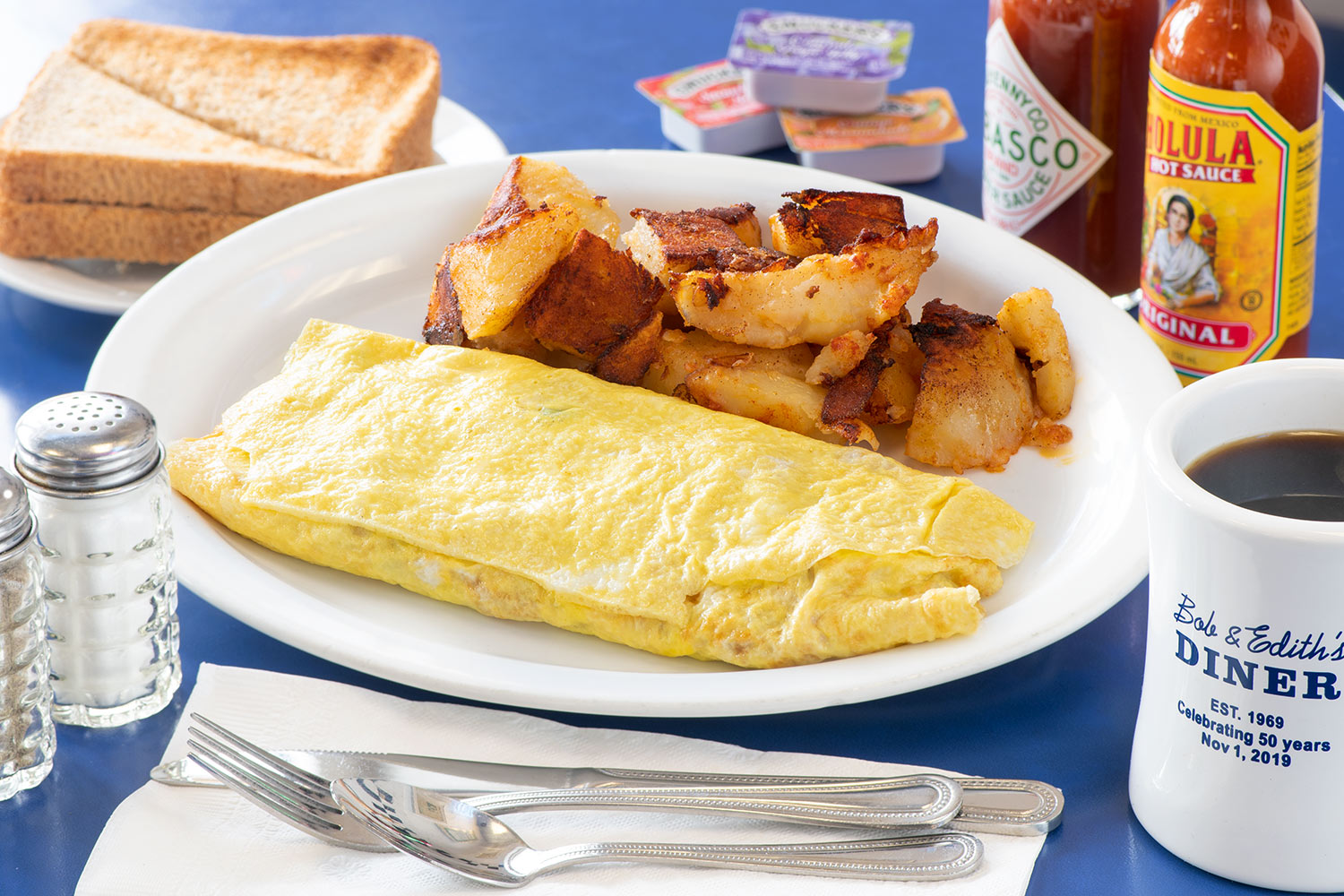 An omelet with home fries in the background, utensils in front, and coffee on the side.