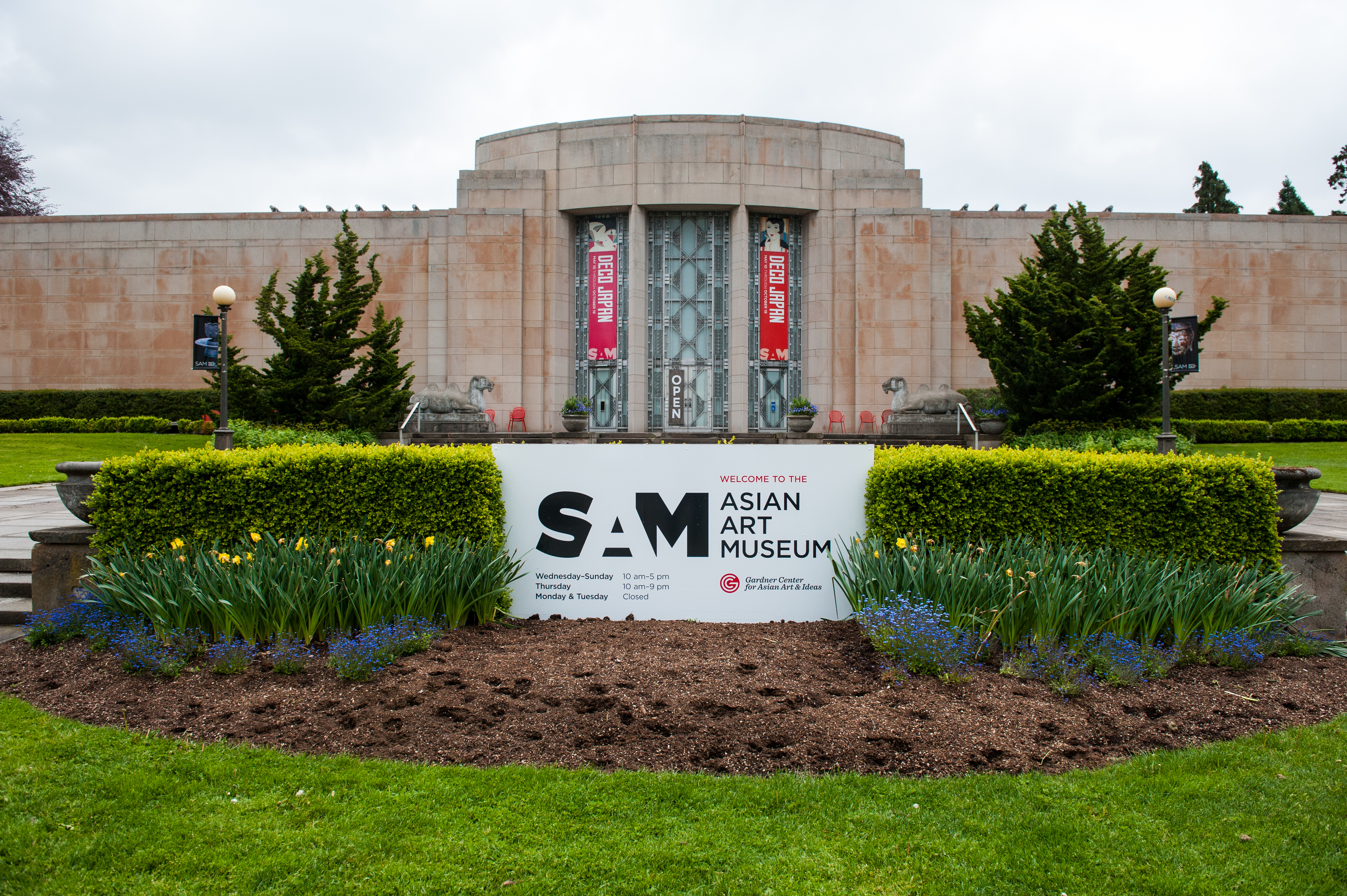 A stone, Art Deco building with a central, rounded bay with three vertical stained-glass windows. There’s landscaping in the front around a sign that says “SAM ASIAN ART MUSEUM.”