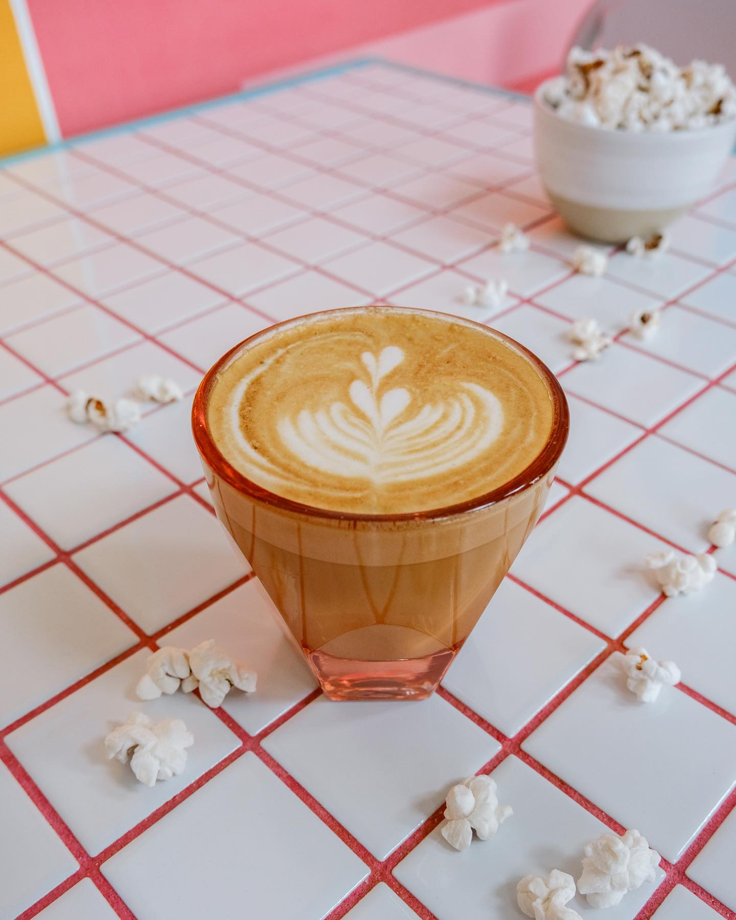A cup of a coffee in a clear red glass with latte art surrounded by popcorn kernels