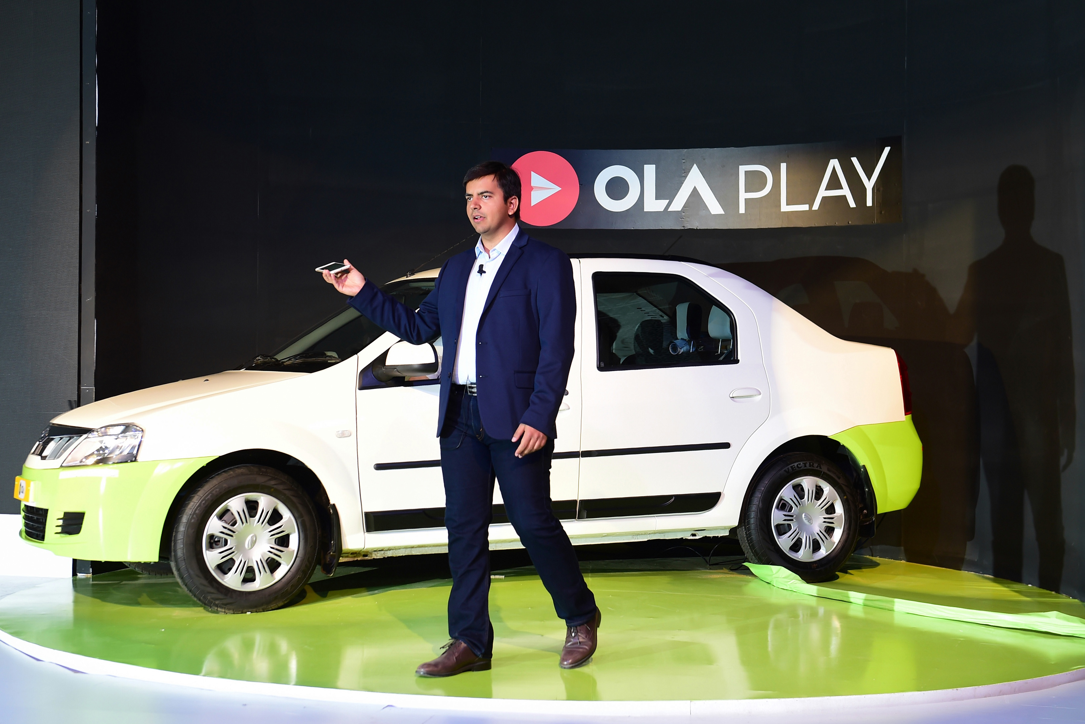 Indian Ola app co-founder and CEO Bhavish Aggarwal stands onstage in front of a car and addresses an audience