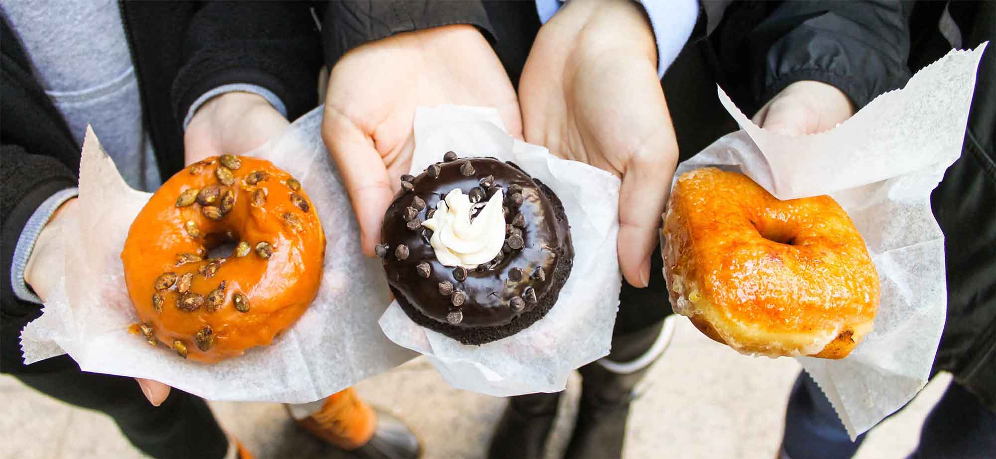 Hands outstretched offering three types of doughnuts, including a seasonal one to the left, a chocolate one in the middle and their creme brulee offering on the right.