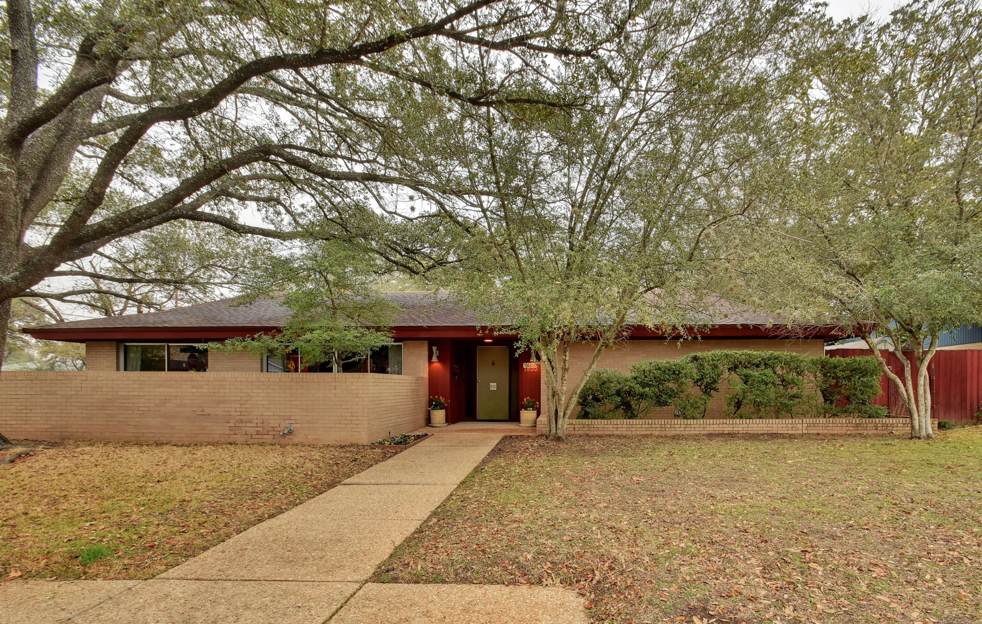Low-slung, large ranch house with off-white brick, walled front courtyard, red trim, big oaks