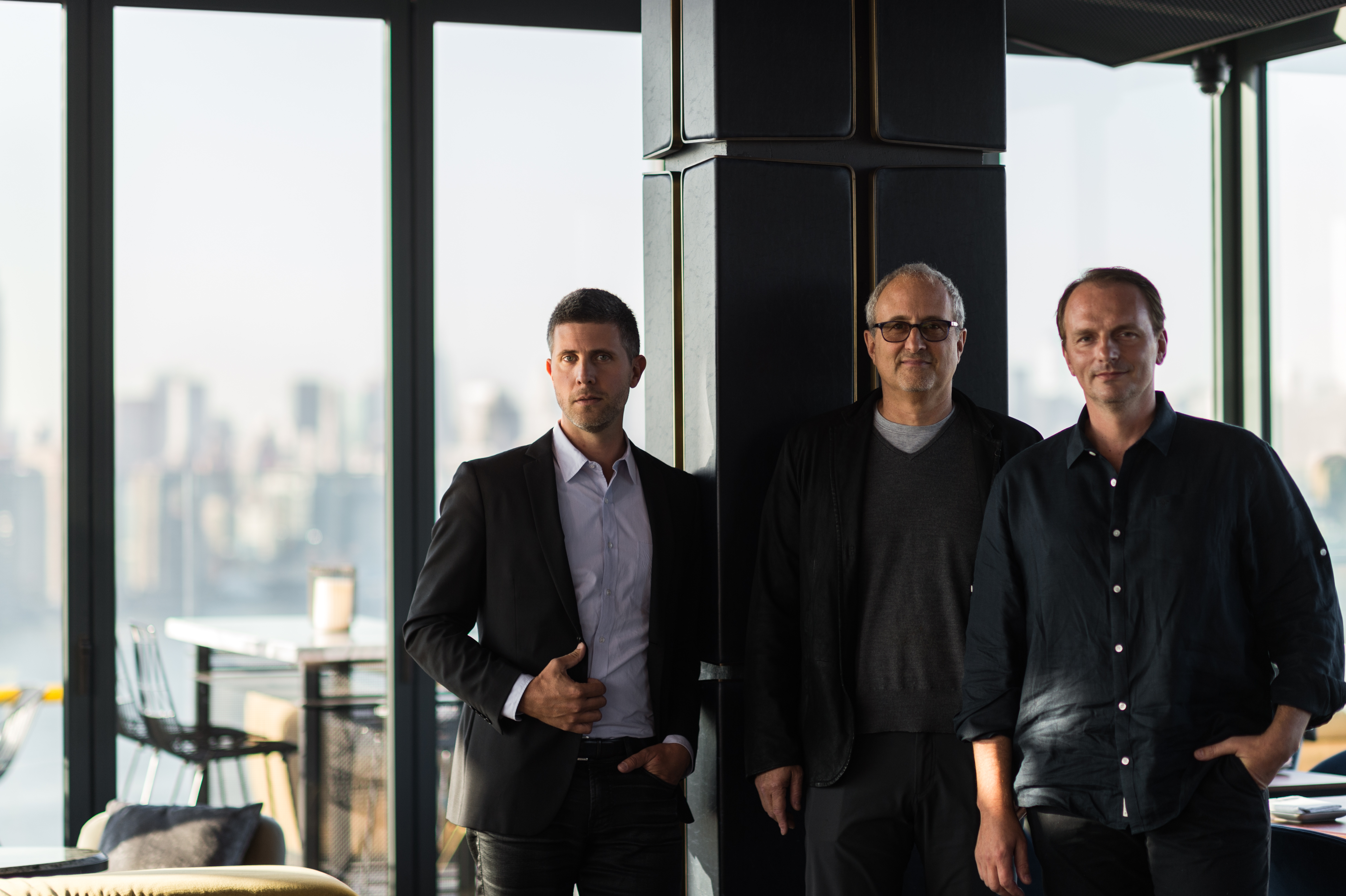 A photo of partners Luke Ostrom, Josh Pickard, and Andrew Carmellini, all dressed in dark colors and standing in front of windows with a view of Manhattan