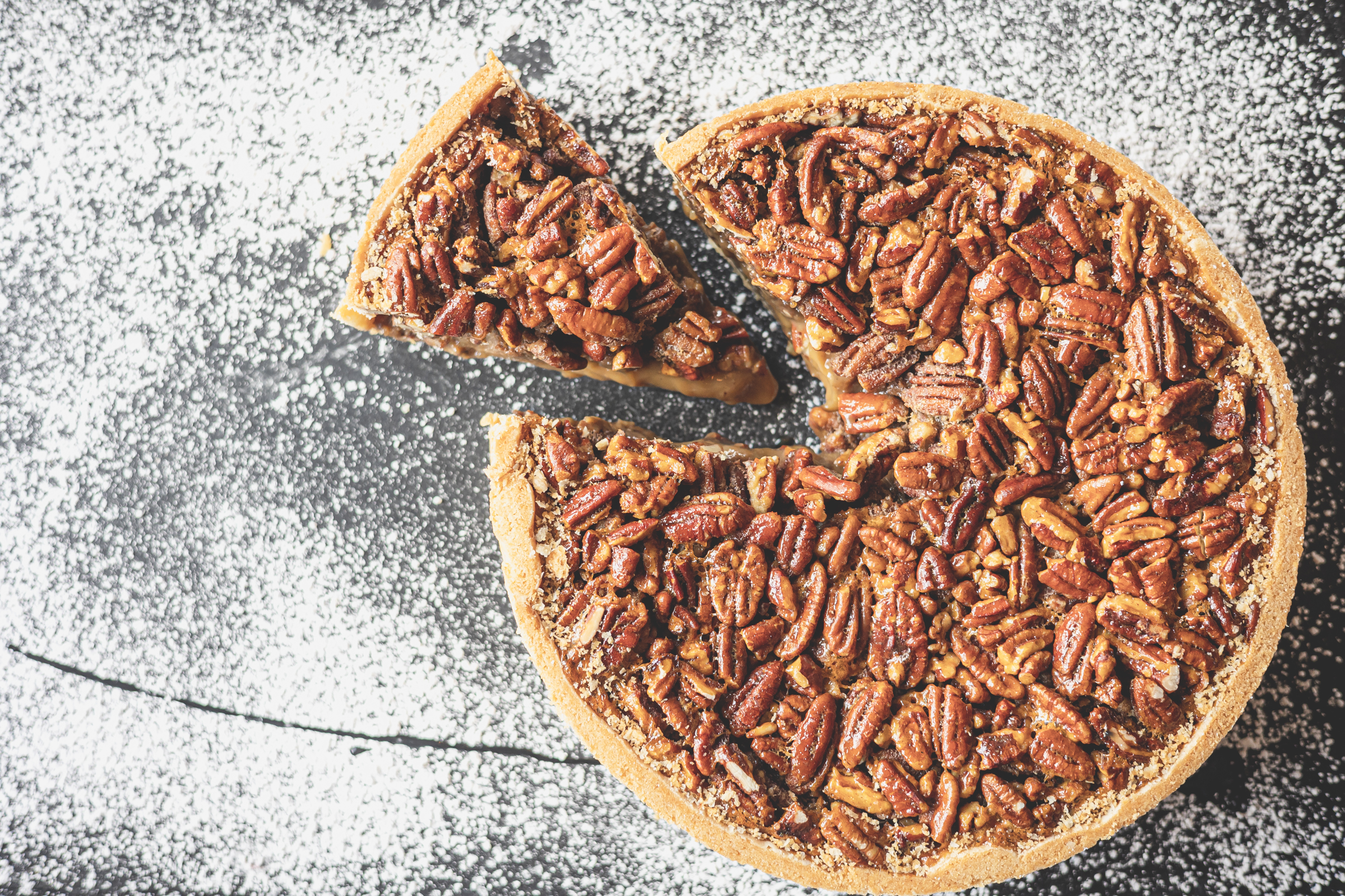 A whole pecan pie with a slice of pie separated, on a table sprinkled with powdered sugar.