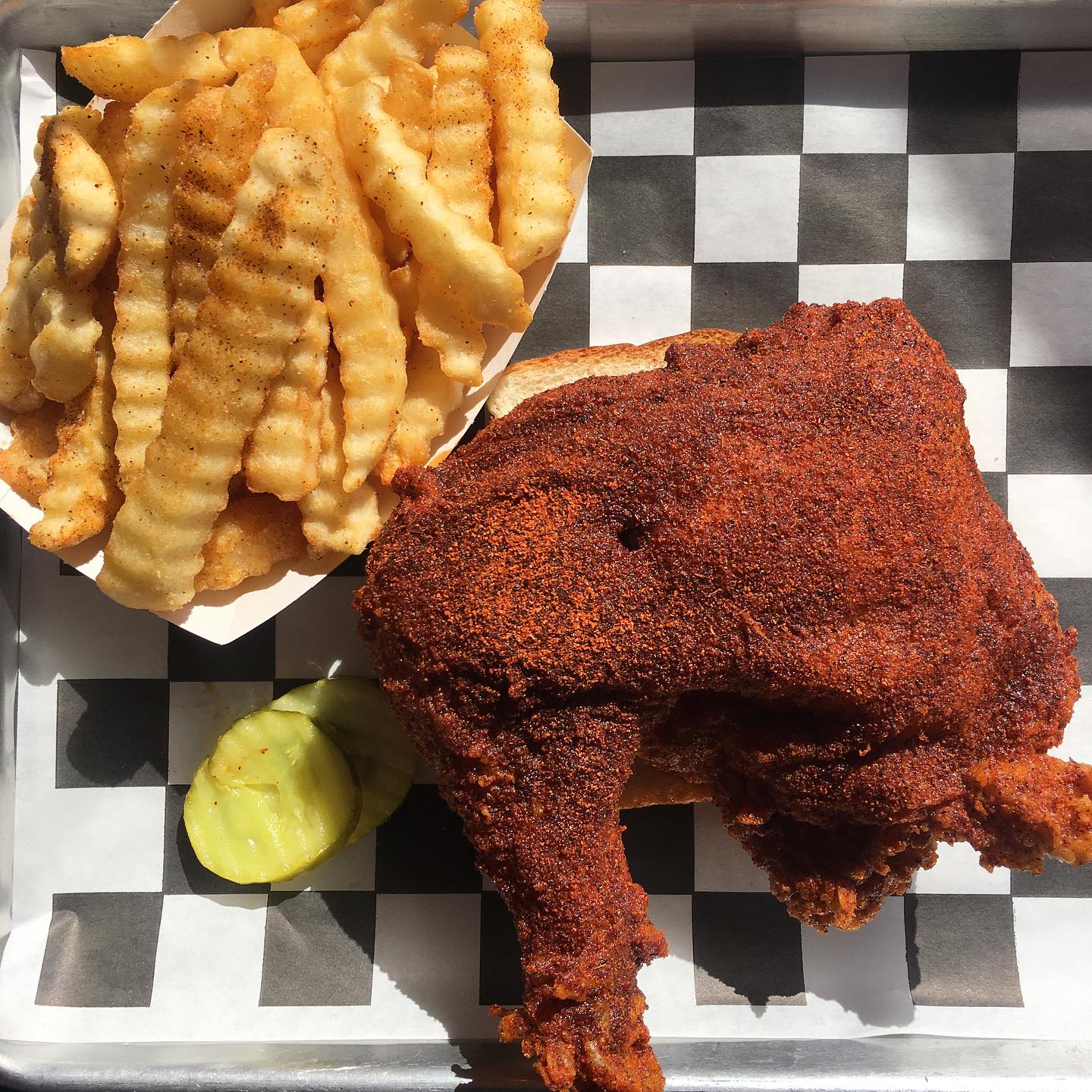 A quarter piece of hot chicken next to a basket of crinkle cut fries. Both items are sitting on a black and white checkered piece of tissue. 