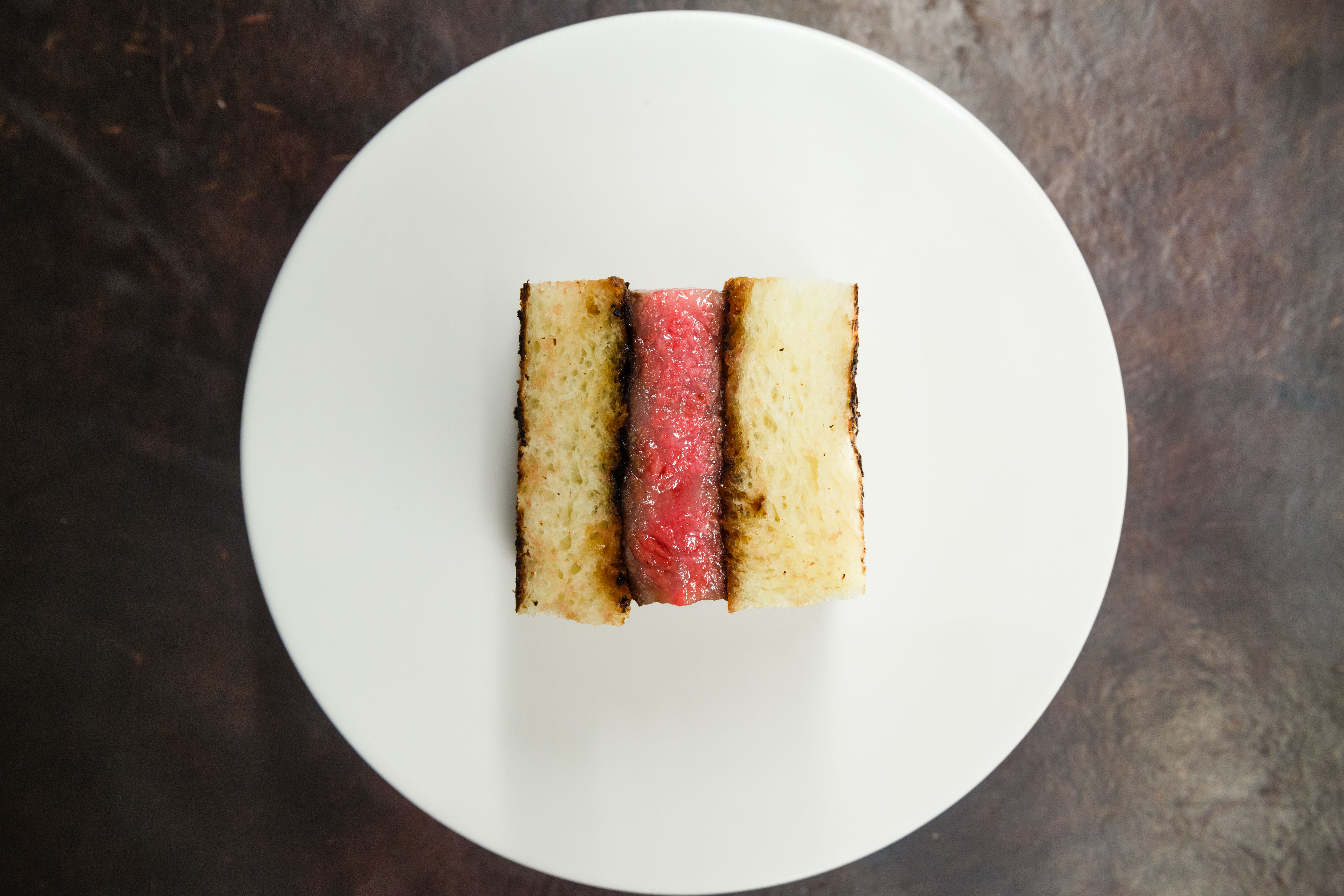 a wagyu sandwich — pink meat between crustless slices of white bread — sits on a white plate.