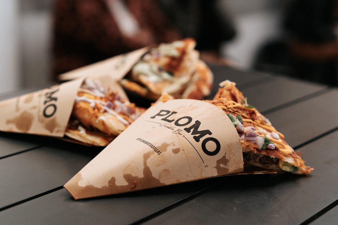 Three quesadillas sit in cardboard sleeves on a wooden table. The sleeves read: “Plomo Quesadillas.”