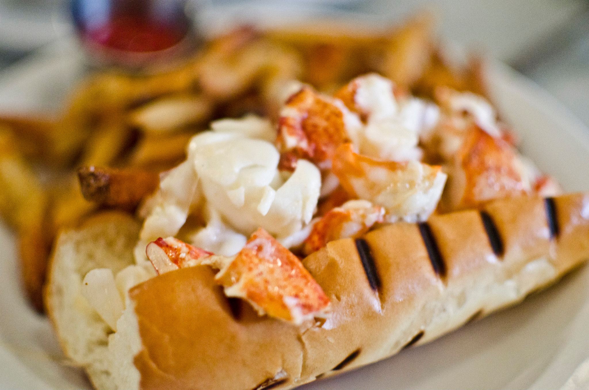 The Maine lobster roll at Neptune Oyster is served on a grilled hot dog bun atop a white plate, and is accompanied by French fries and a ramekin of ketchup. 