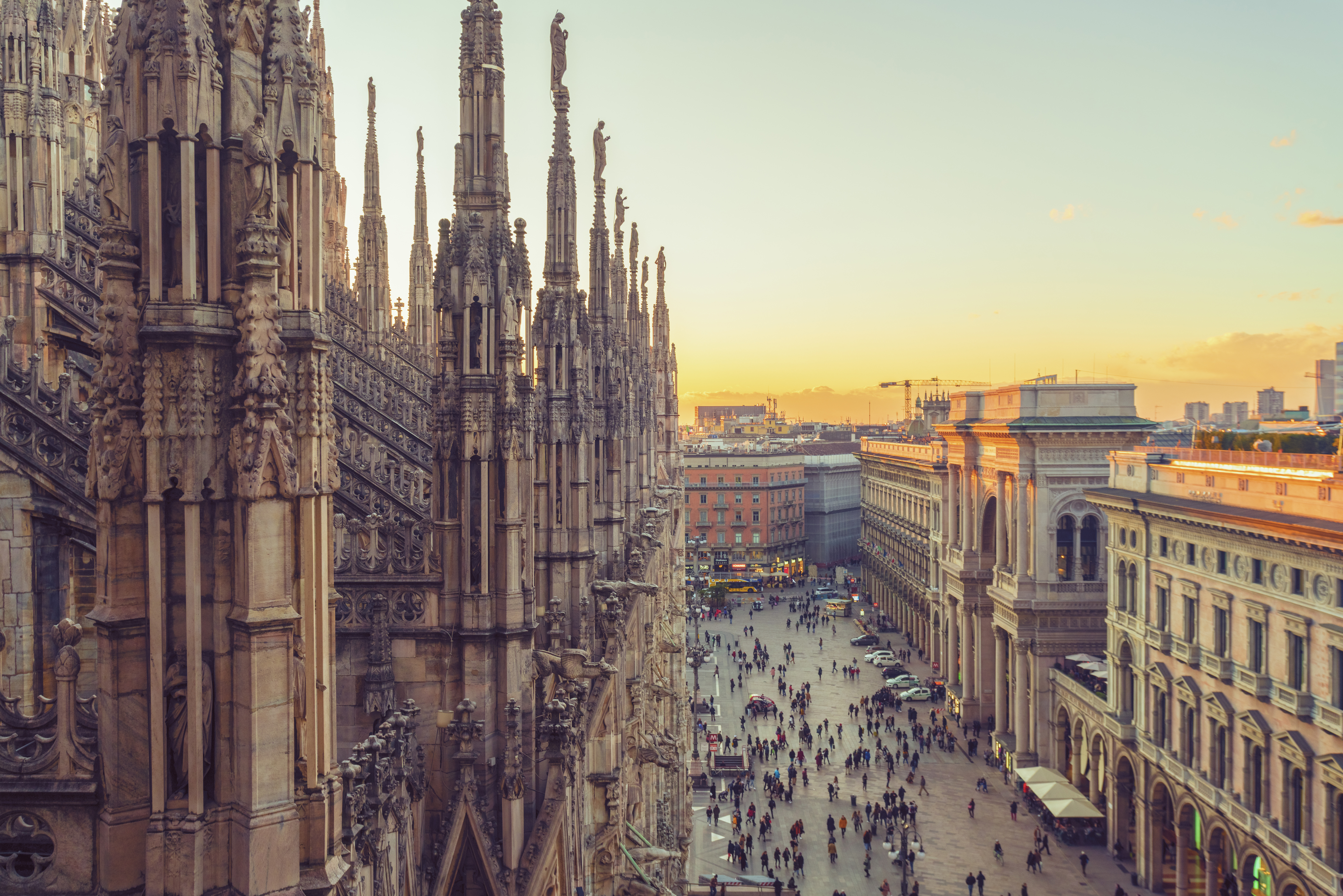 An aerial view of Milan, Italy. There is a building with ornate architecture in the foreground. Below there is a plaza with many people. Surrounding the plaza are various city buildings. 