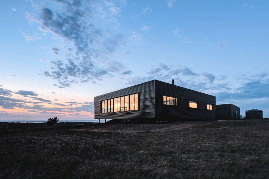 Cube-like home with black-clad exterior on rocky coastal terrain. 
