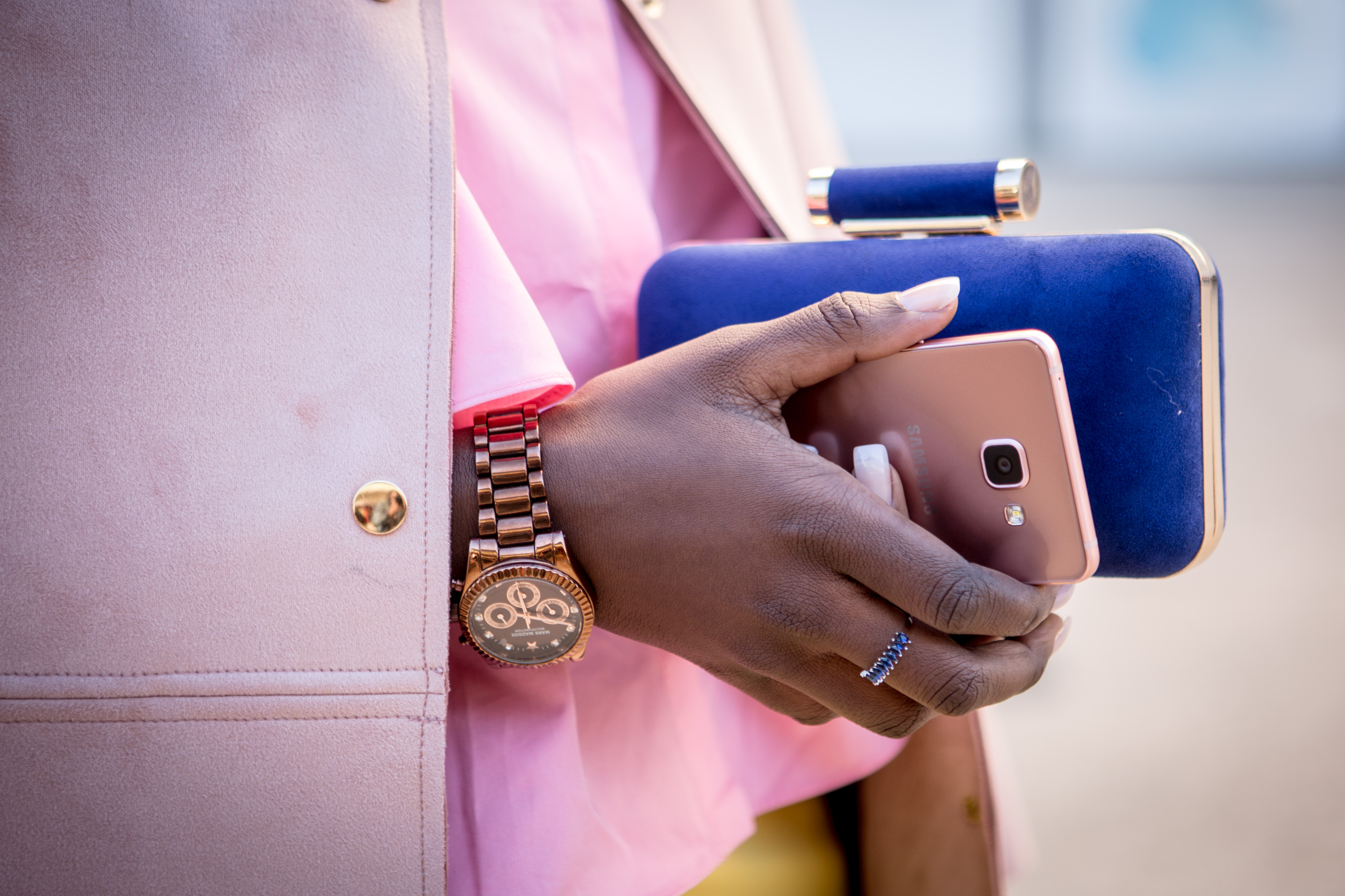 A close up of a woman wearing a pink jacket and a gold watch, carrying a rose gold smartphone and a blue purse.