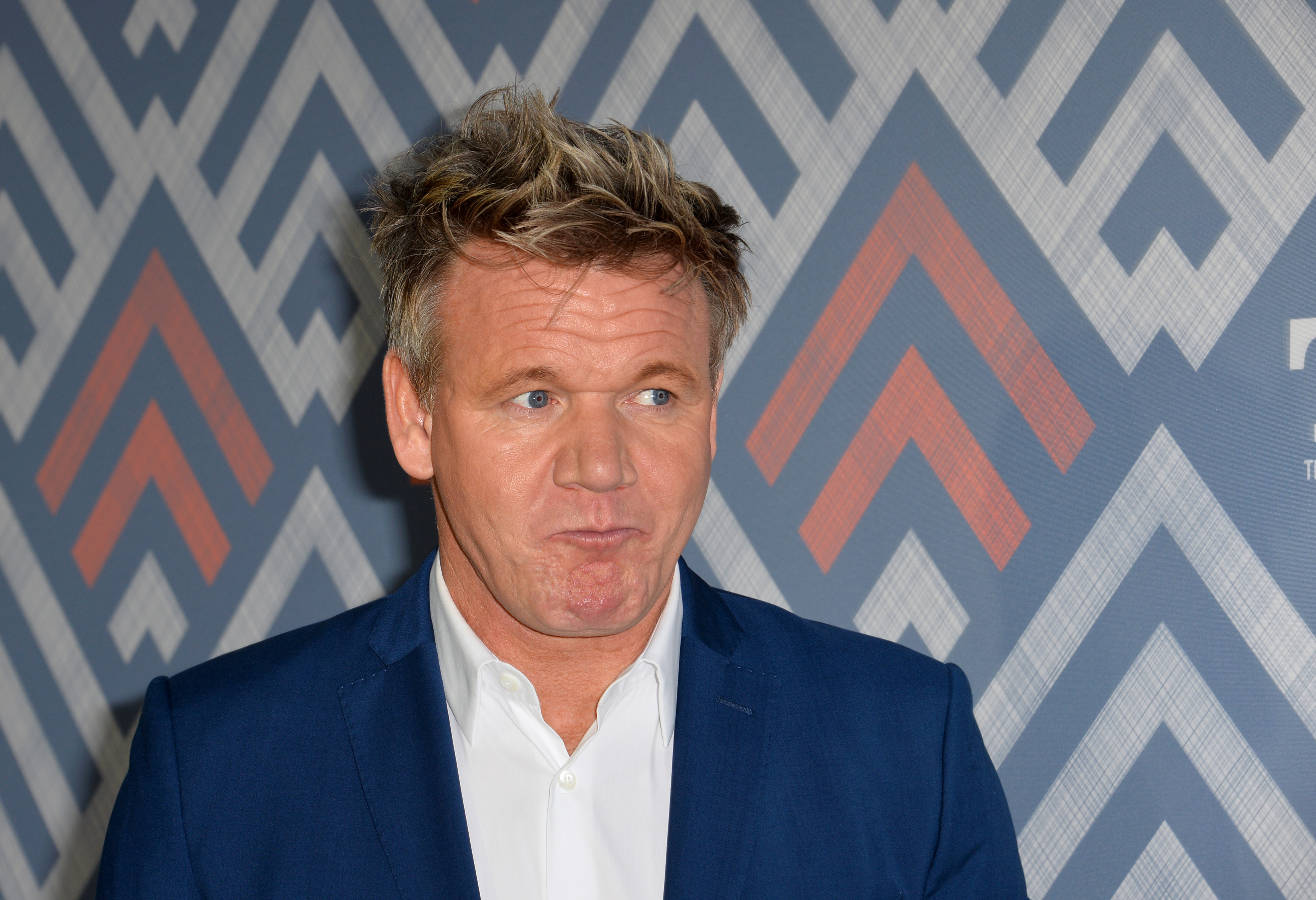 Gordon Ramsay wears a suit against a chevron-printed background, while pulling a strange face