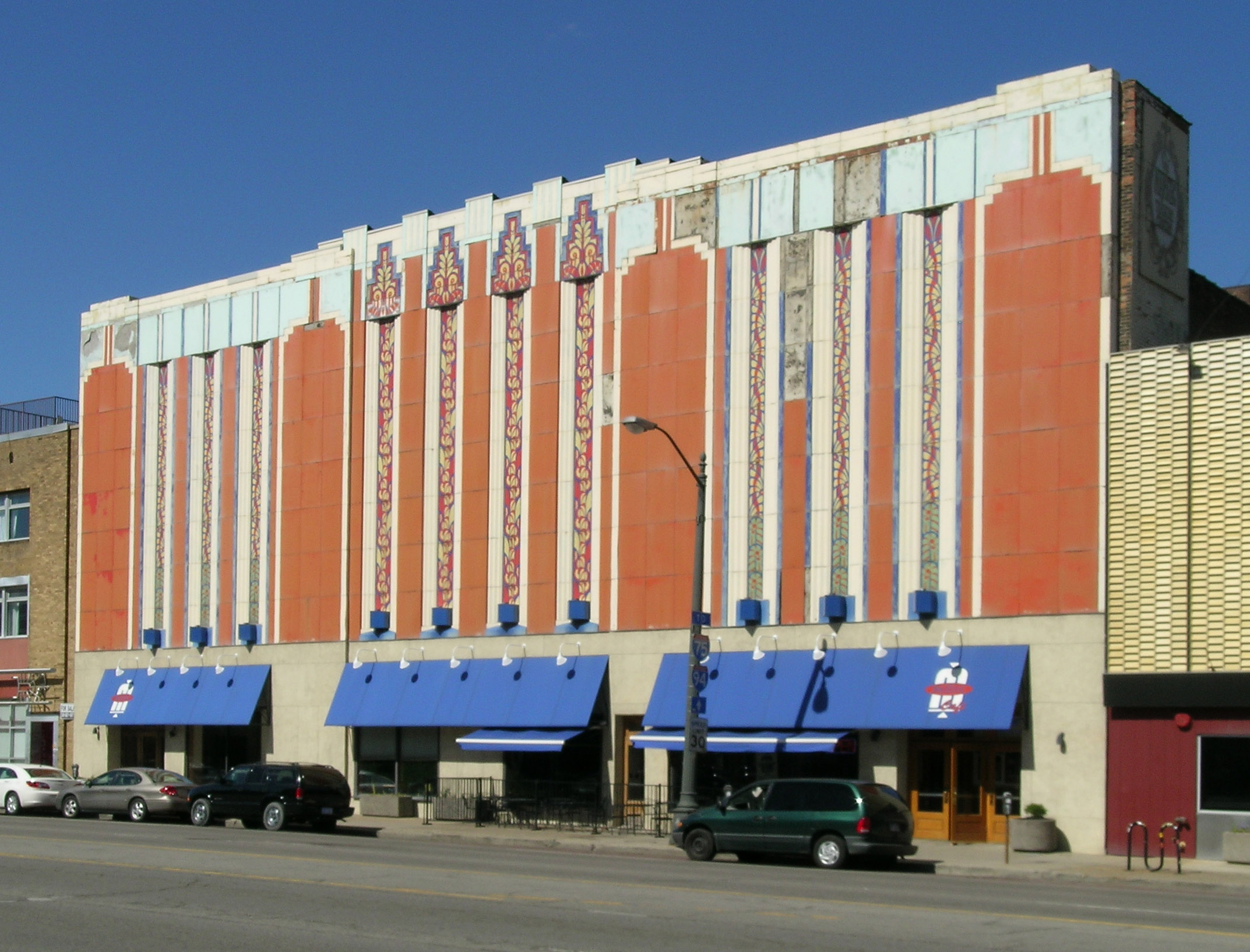 Facade of a several-story Art Deco building with orange paint and vertical strikes of blue and white. There’s also a blue awning over the ground-floor windows.
