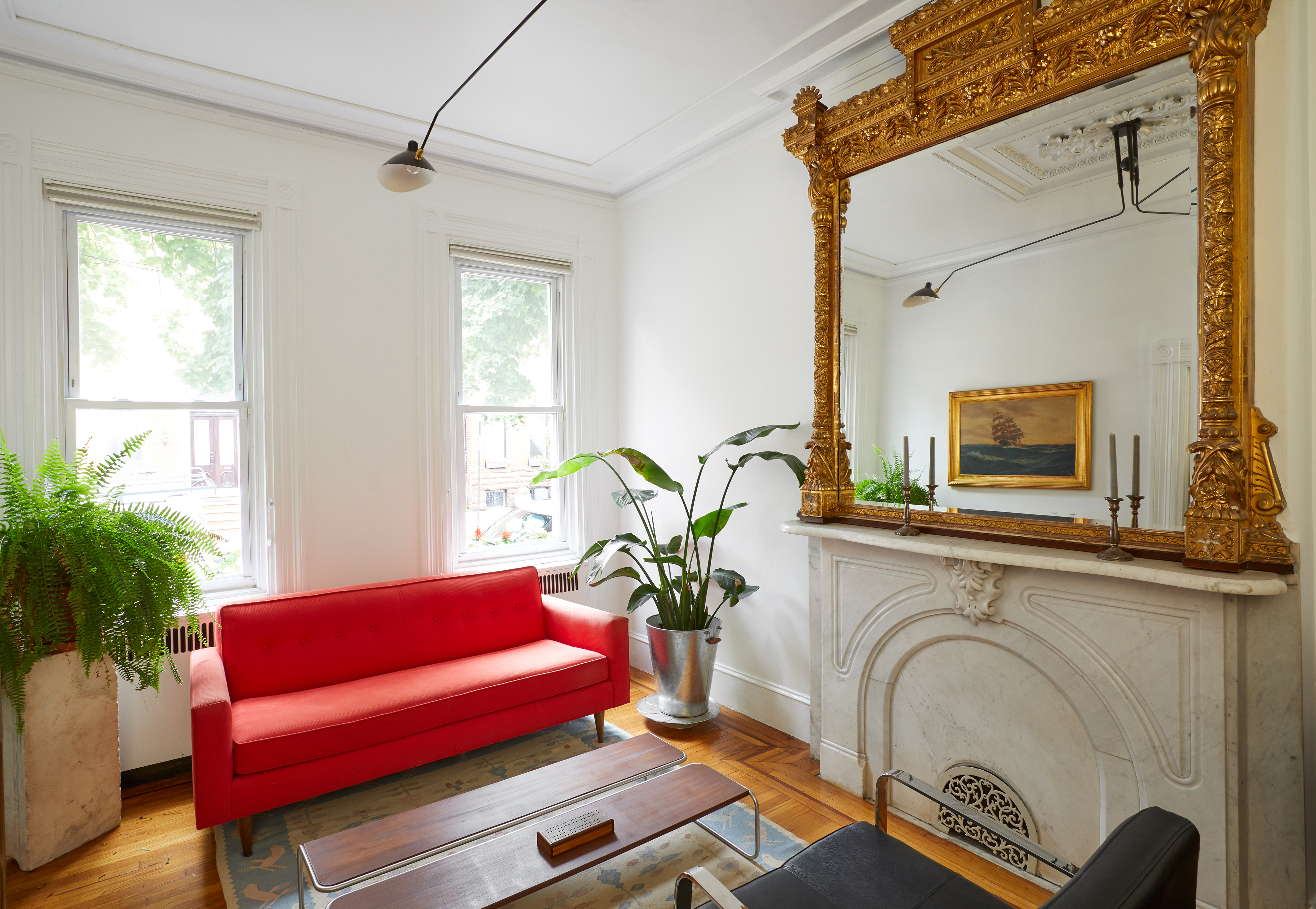 A living area. There is a bright red couch and a fireplace. Above the fireplace sits a large mirror with an ornamental brass frame. There are two windows letting in natural light. There are houseplants in planters on the floor and a wooden coffee table. 