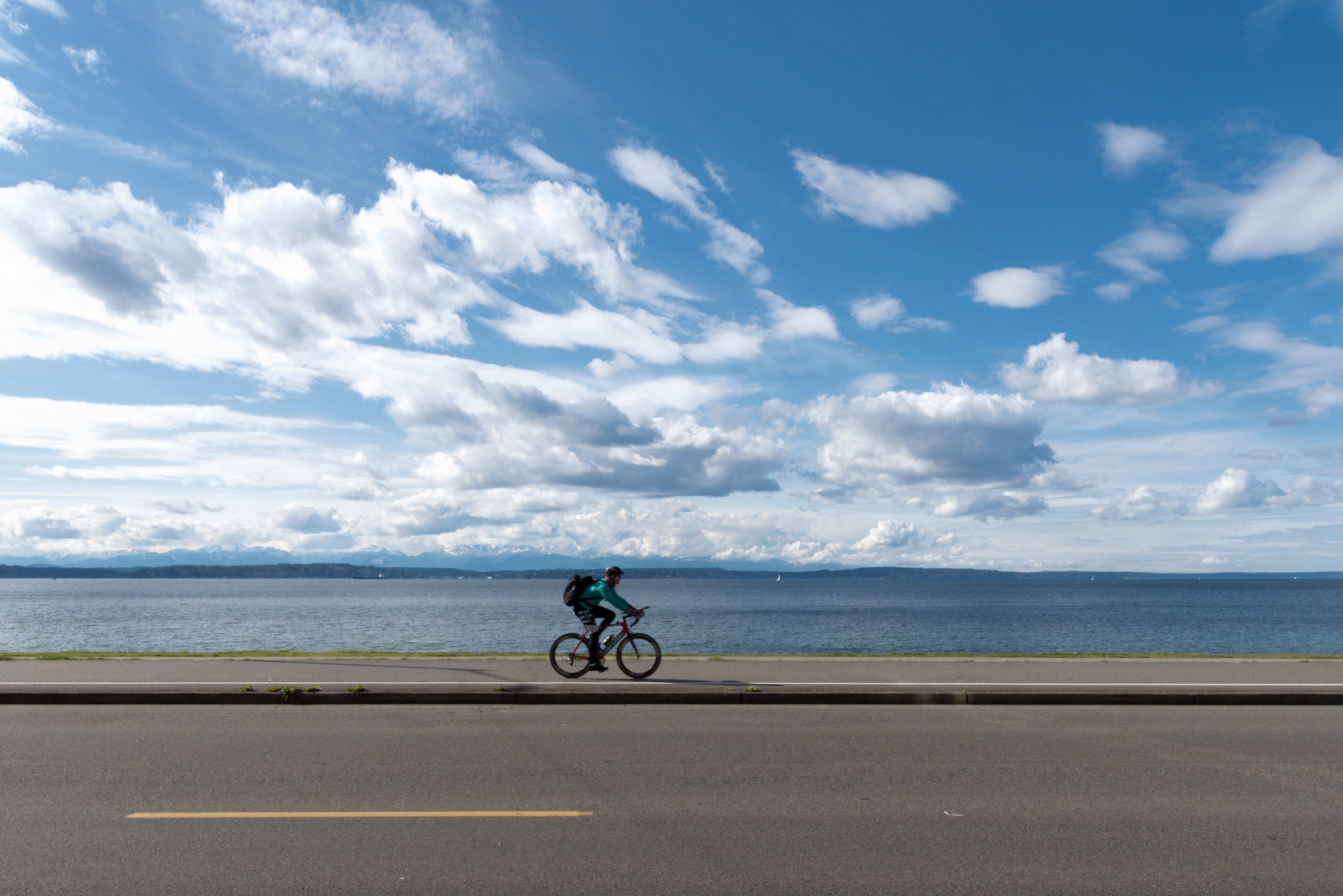 In the foreground is a highway with a bike lane. There is a person riding a bicycle in the bike lane. In the distance is a body of water and a sky with many white clouds. 