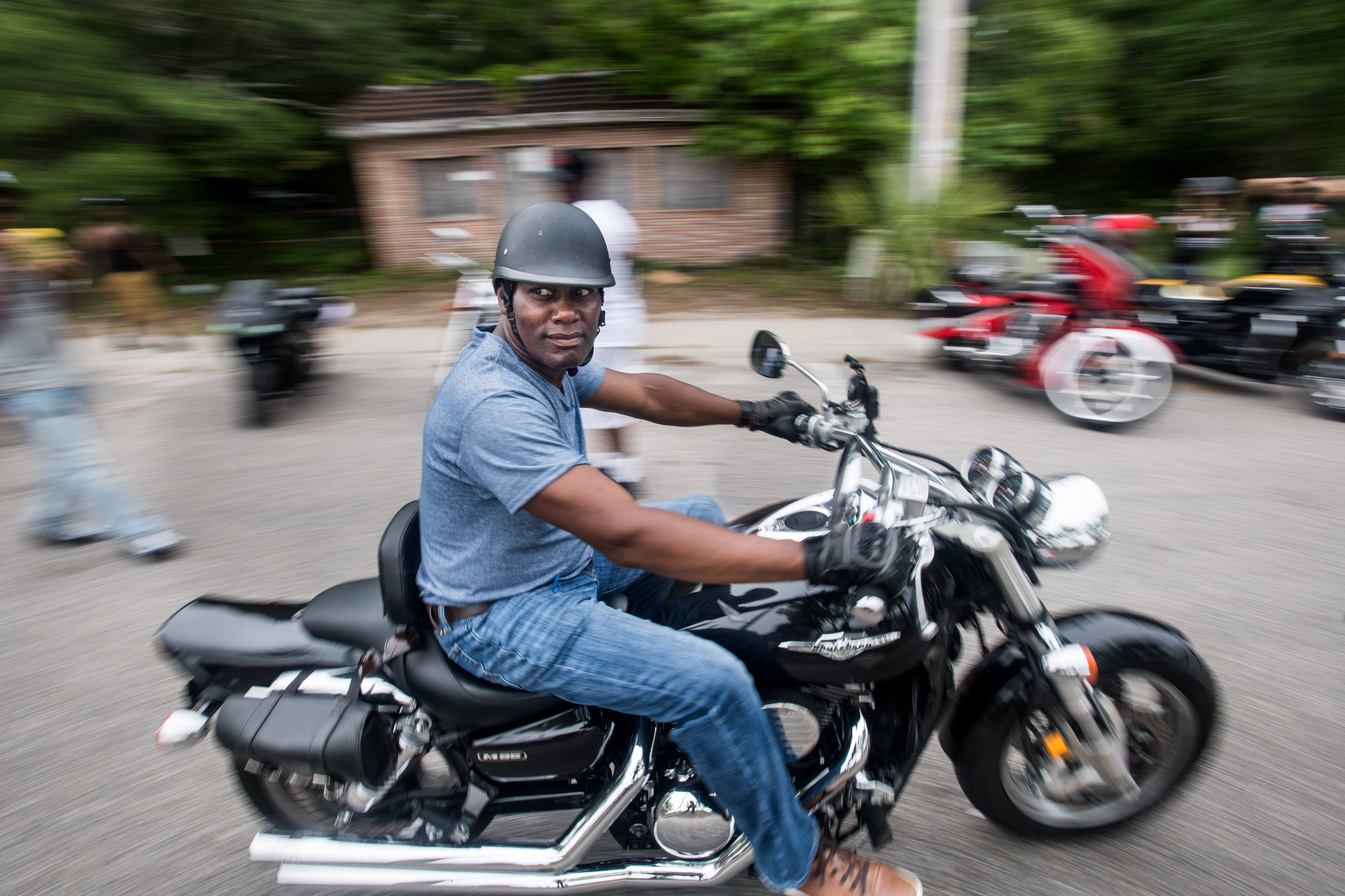 A black man in jeans and a minimalist motorcycle helmet rides a large black motorcycle during Black Bikers Week.