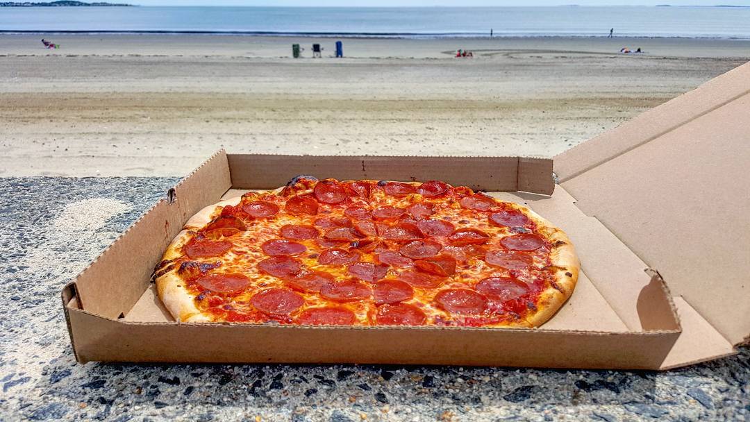 A pepperoni pizza sits in an open cardboard pizza box on a stone surface, with a mostly empty beach visible in the background
