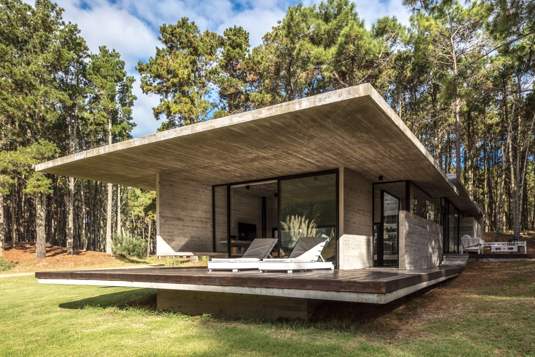 This summer home sits on a slight slope in a clearing among pine and acacia trees and is built from board-formed concrete and large glazed windows. Overhanging concrete slab roofs provide a sense of privacy, while a terrace and a deck lends themselves to