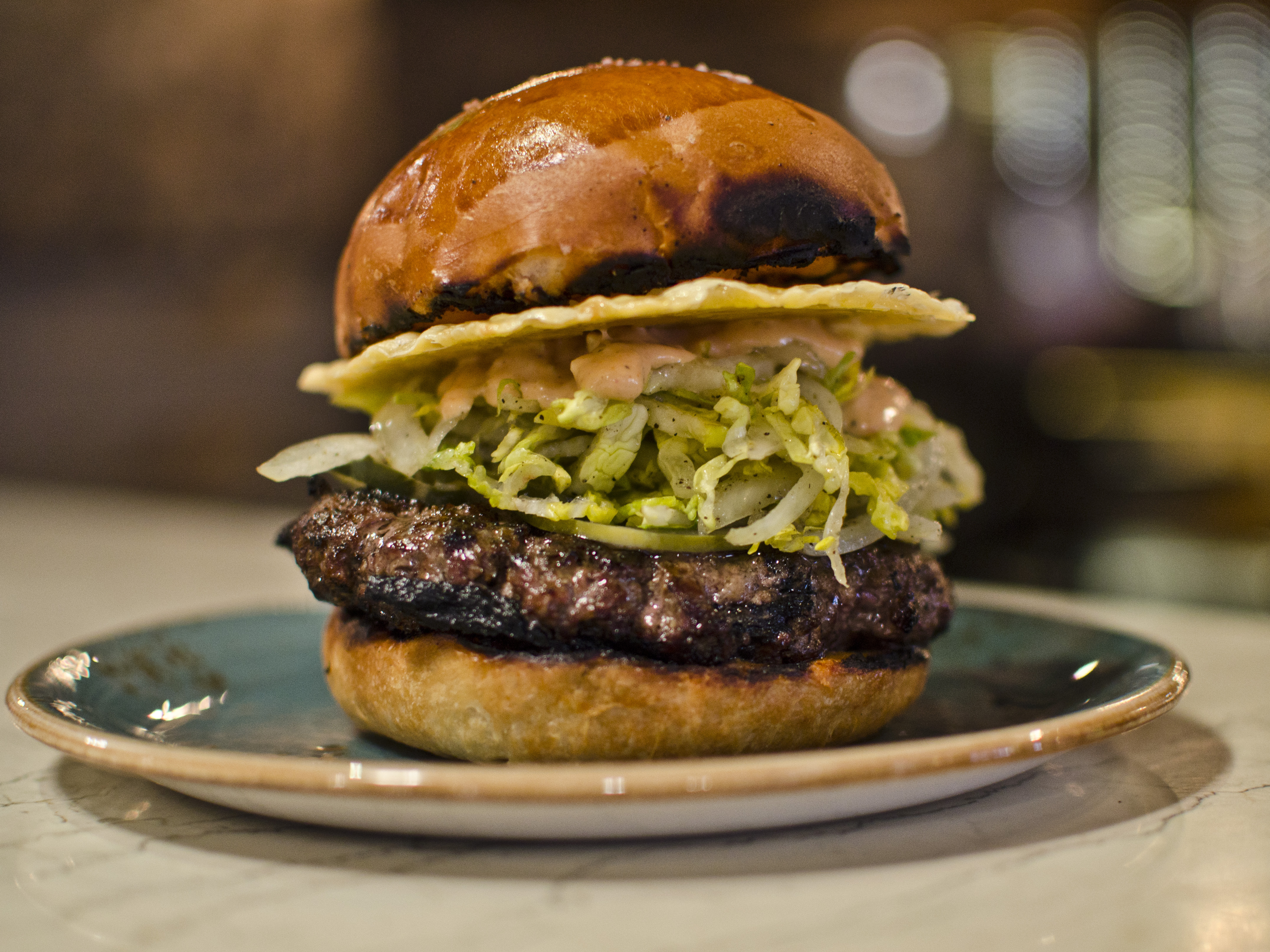 A burger sits on a blue plate, piled high with shredded lettuce, sauce, and a crispy disc of cheese