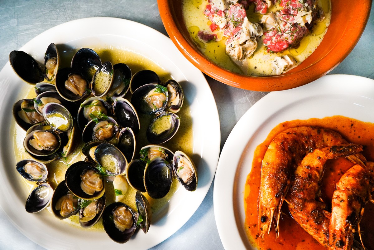 Clams with vinho verde sit on a white plate next to large, head-on shrimp on a separate plate in this overhead shot.