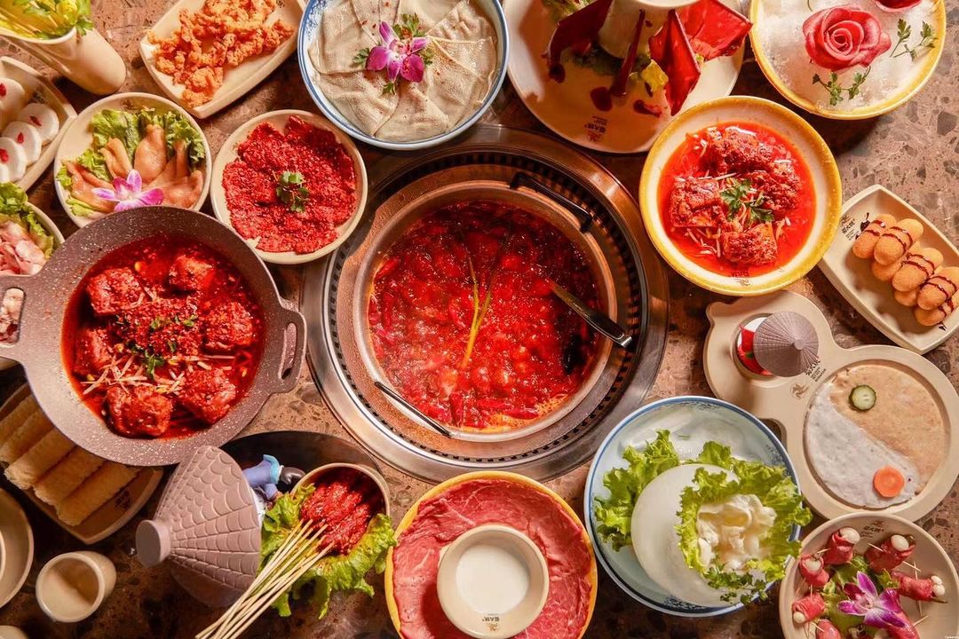 Overhead view of a table full of Chinese hot pot plates, including a fiery red broth, raw vegetables, raw meats, and more.