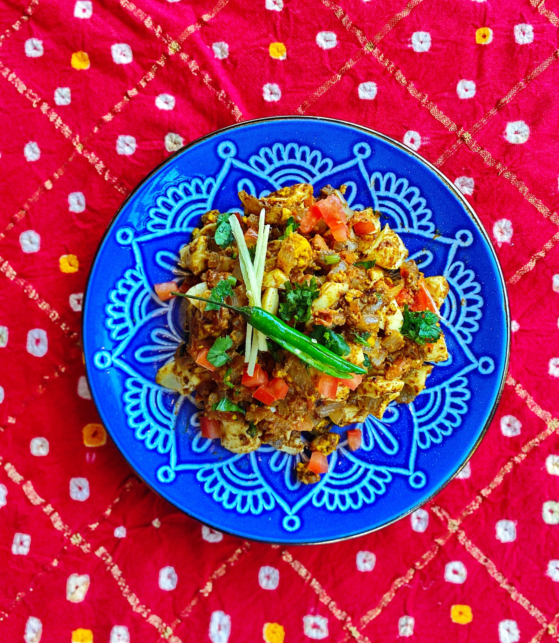 Shredded egg tossed with veggies and southern Indian spices on a bright blue plate sitting on a red pattern tablecloth.