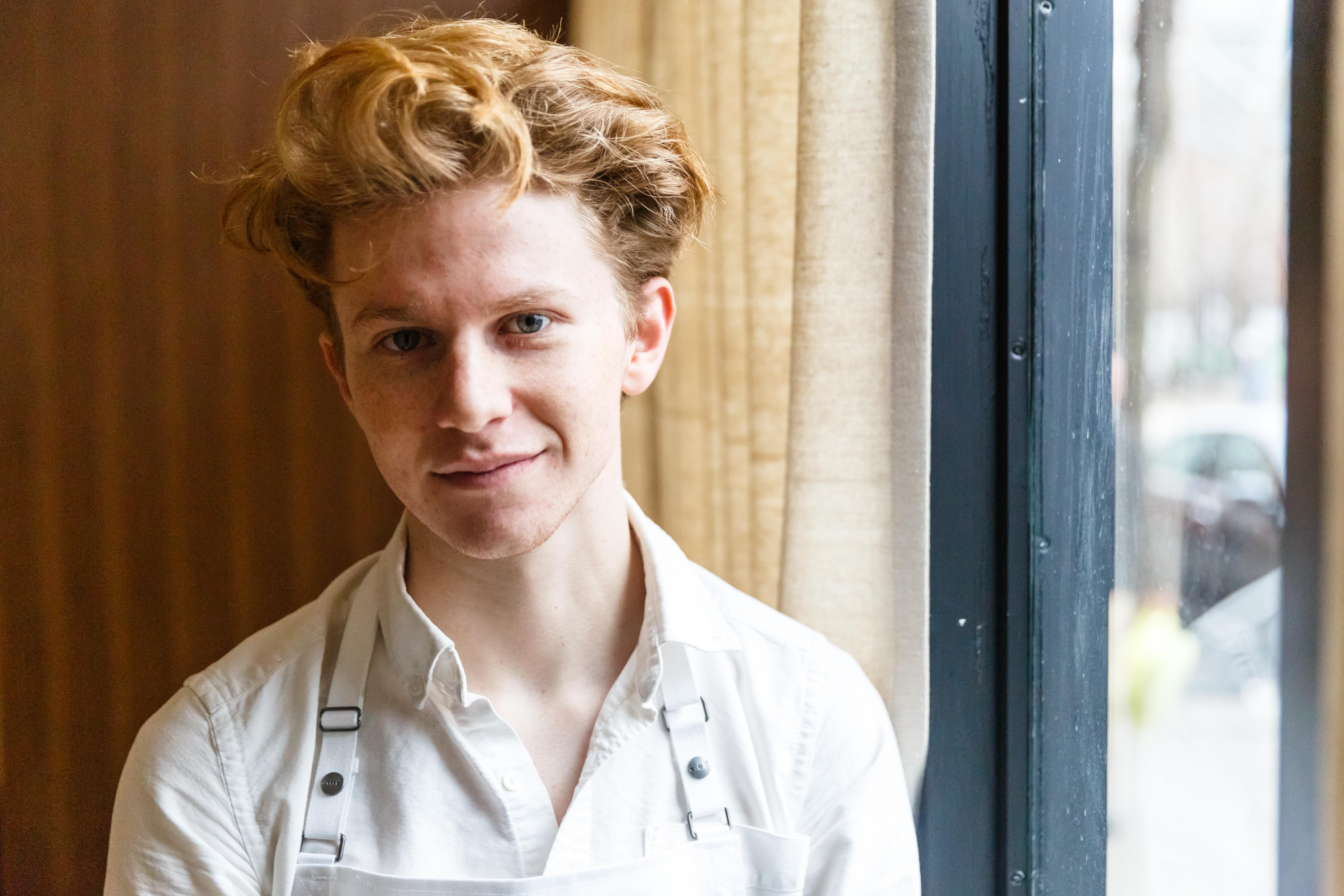 A young person wearing an apron and chef’s whites, Flynn McGarry, stands in the window of a restaurant.